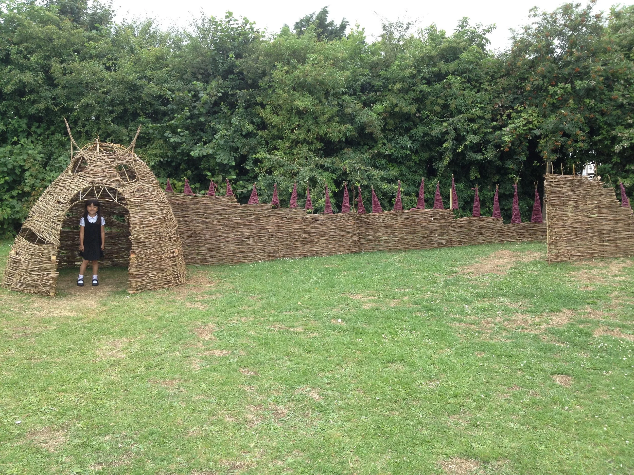 Children’s play area made of woven branches and twigs, resembling a castle or fort, with a girl standing underneath an arch entrance.