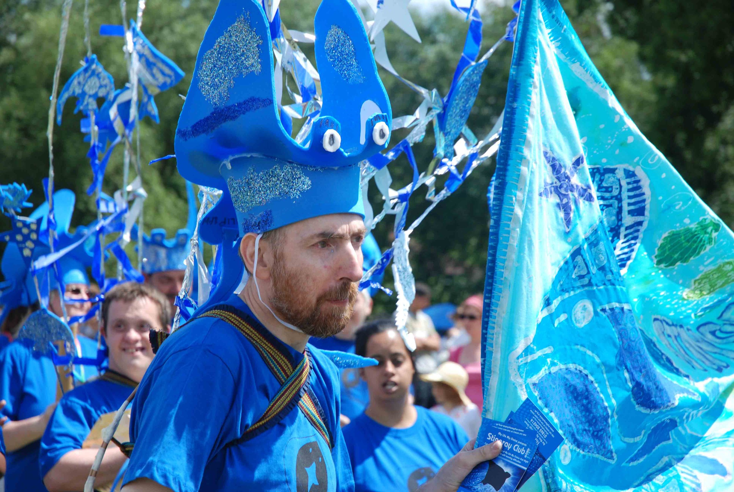 A group of people dressed in blue, participating in a parade or festival, with some individuals wearing handmade blue bunny ears and face paint, holding blue flags and balloons.