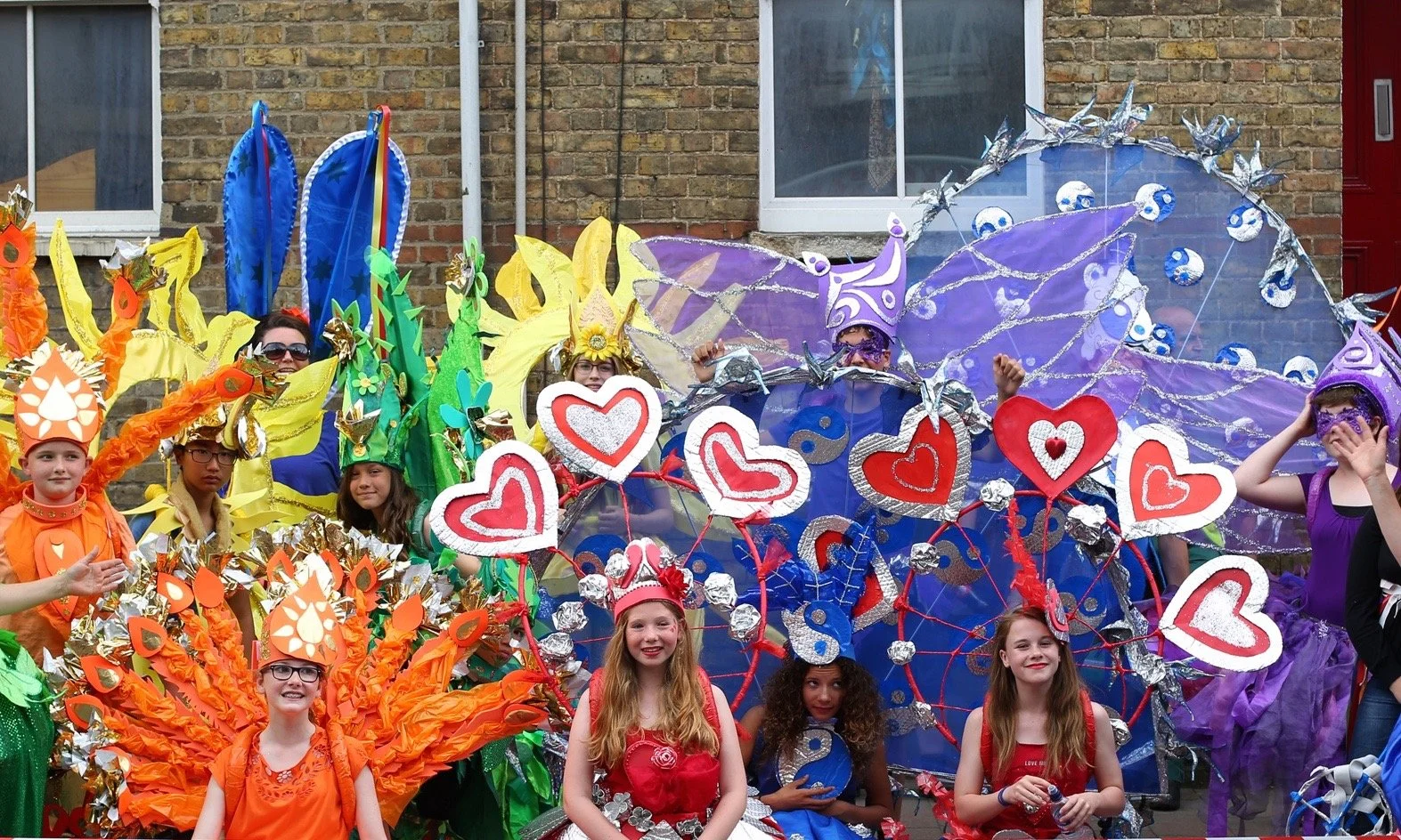 Children dressed in colorful costumes participating in a parade or festival, with some wearing butterfly and fairy costume wings and headpieces, and others holding decorated hearts and flowers.