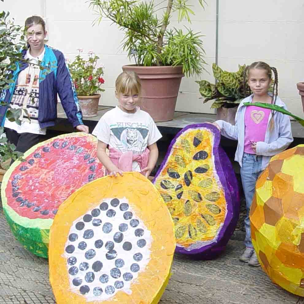 Three children standing outdoors with large, colorful, paper mache fruit sculptures, including a watermelon, pineapple, papaya, and mango, with potted plants in the background.