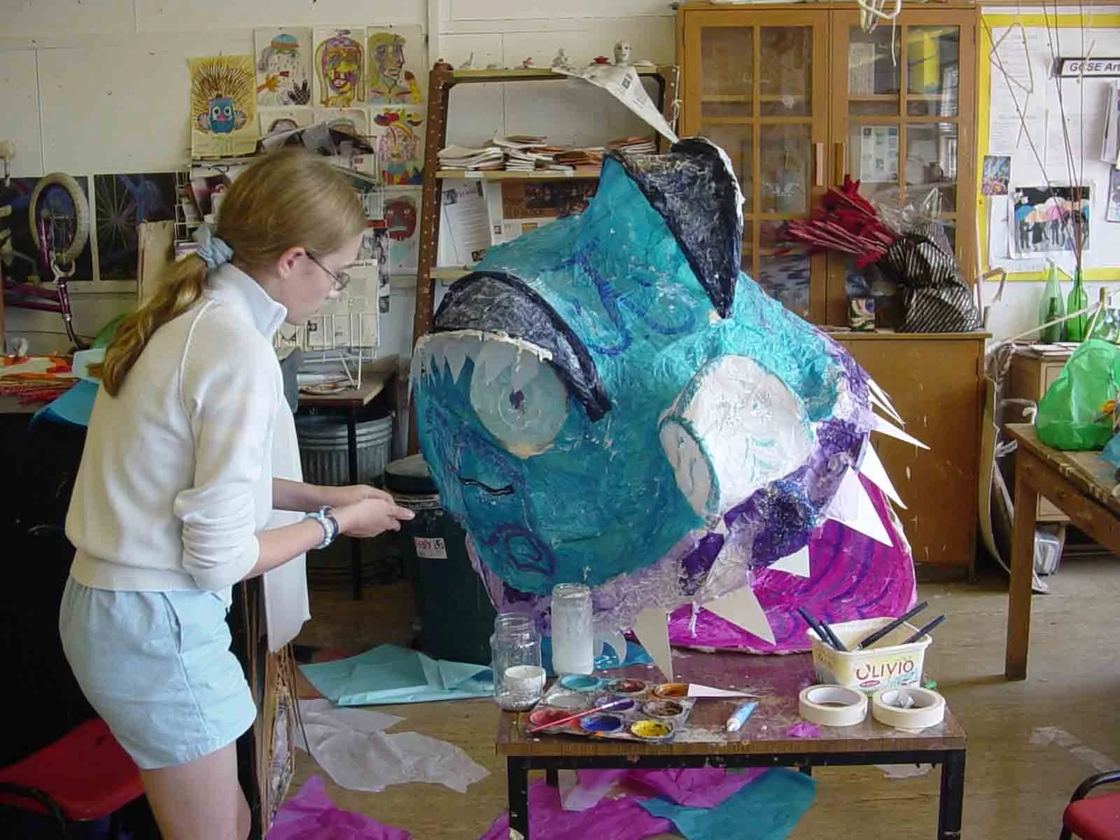 A woman working on a large, colorful paper lantern shaped like a fish, in an art studio filled with art supplies, paintings, and crafts.