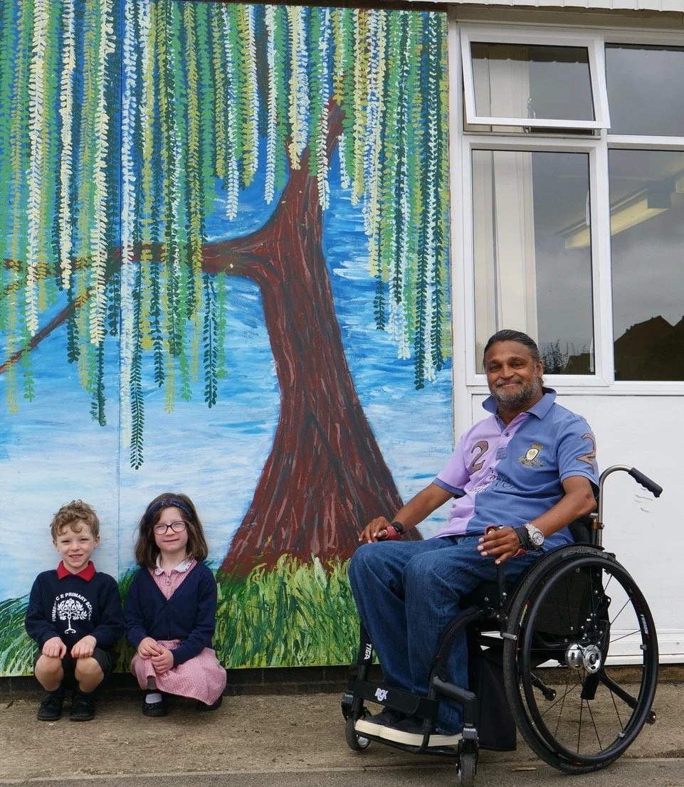 A man in a wheelchair with a beard and two children, a boy and a girl, kneeling on the ground in front of a colorful painted mural of a tree with green leaves hanging down, on an outdoor wall next to a building.