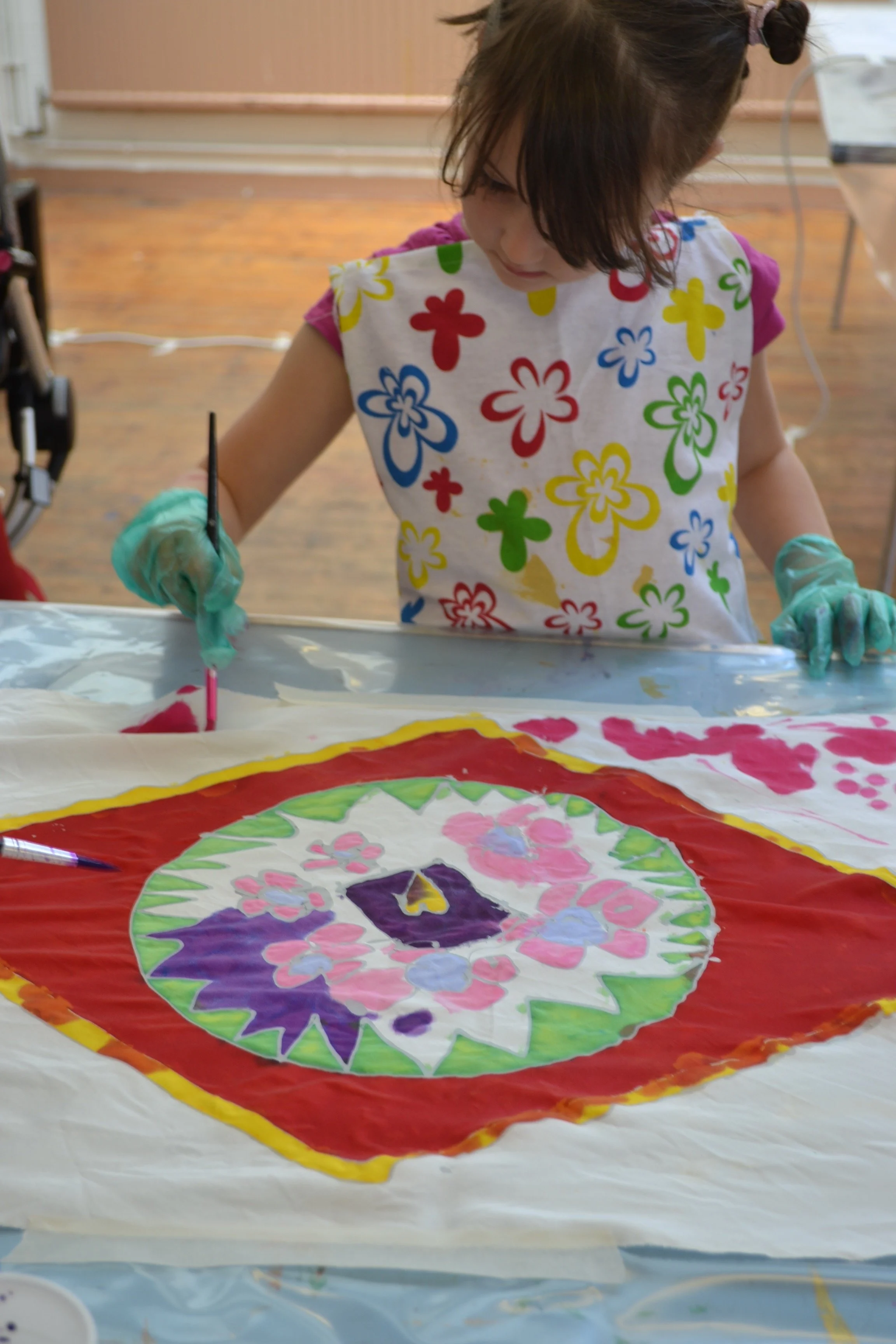 A young girl wearing a colorful flower-patterned apron and gloves is painting a large, circular canvas with various pink, purple, and green floral designs on a table.