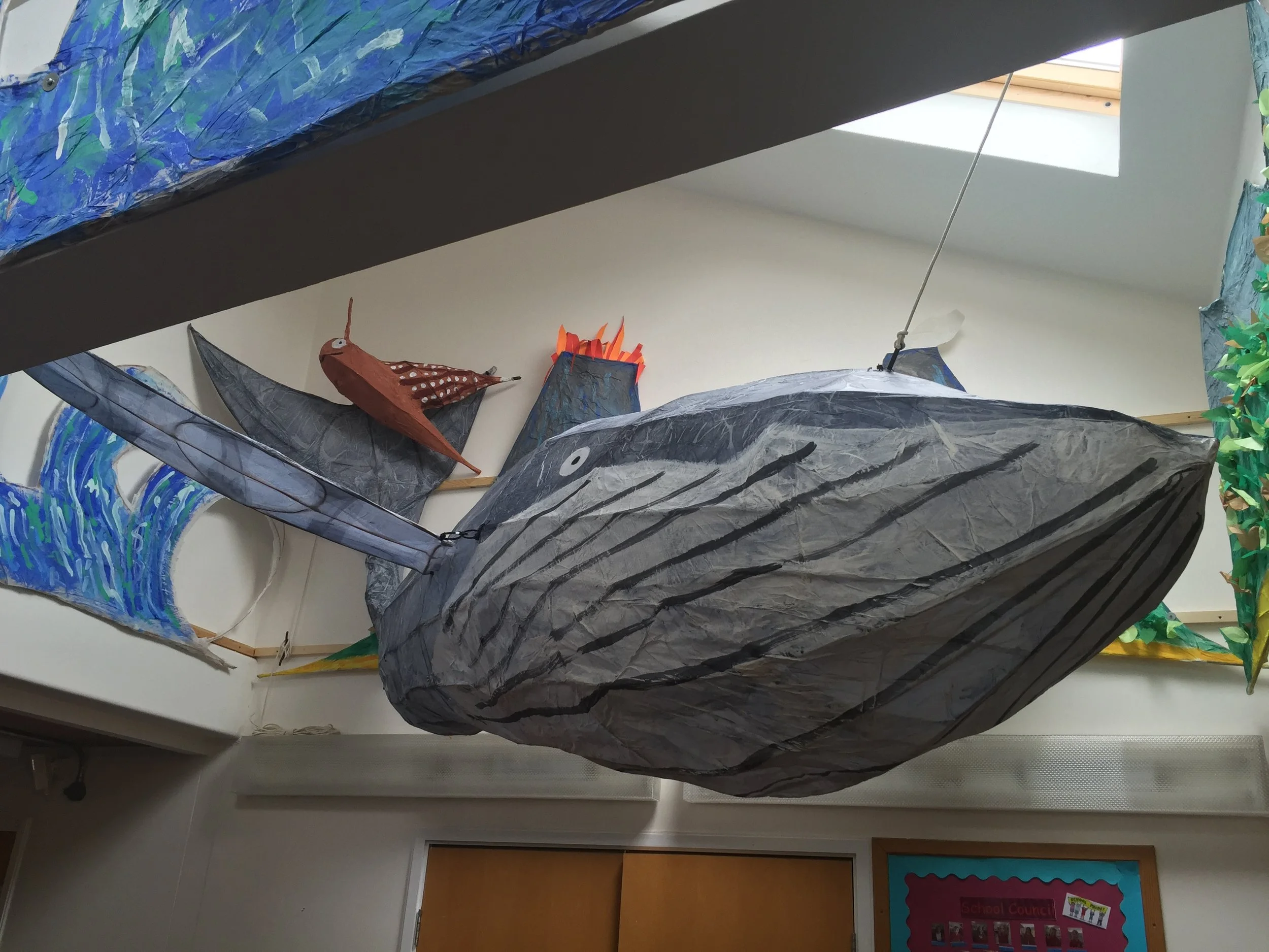 Various colorful, large paper-mounted fish shaped art pieces hanging from the ceiling in an indoor space, with a skylight in the background.