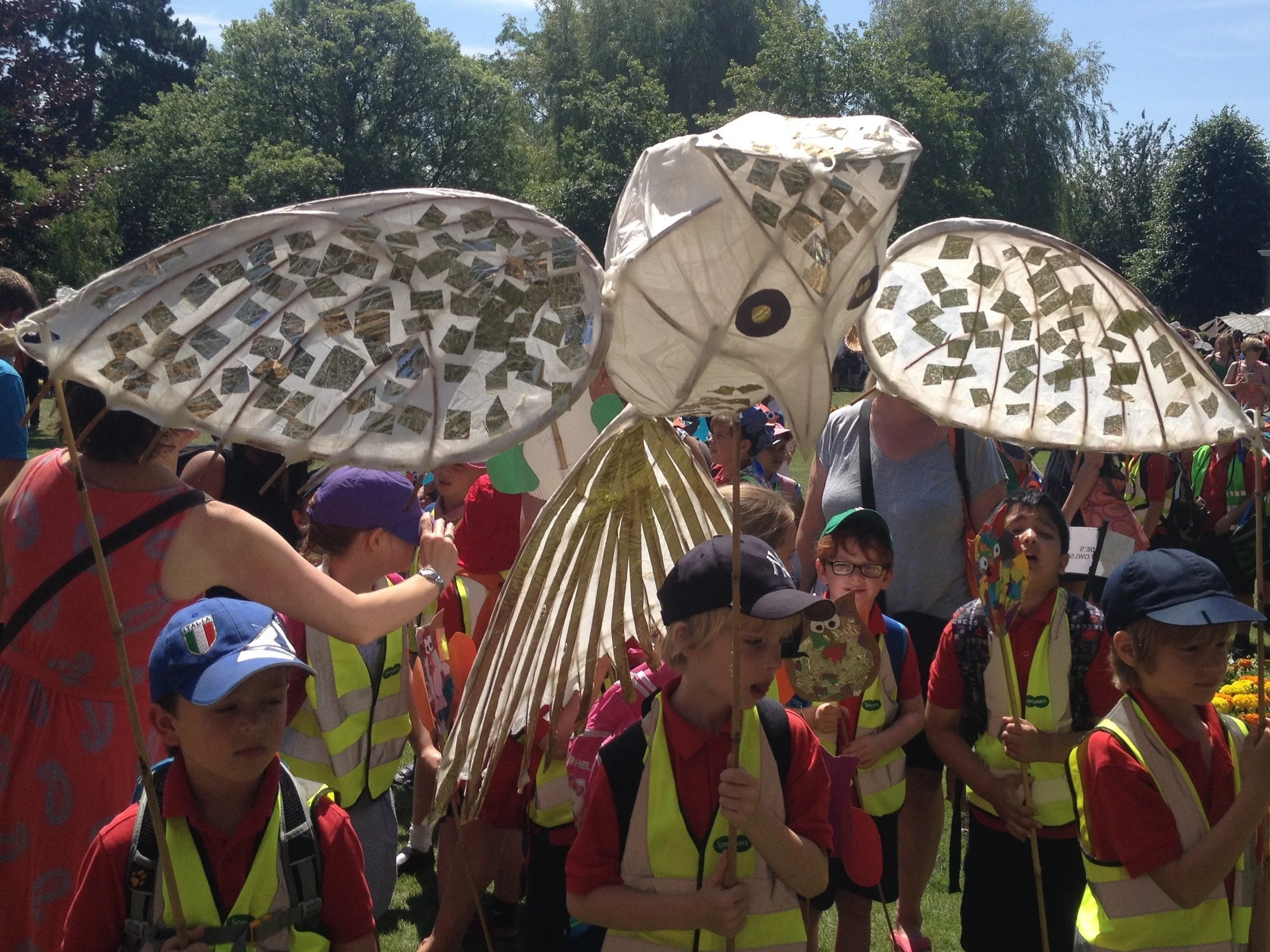 A group of children and adults gathered outdoors in sunny weather, holding colorful paper parasols, some decorated with pictures of animals and nature, with trees and a clear blue sky in the background.