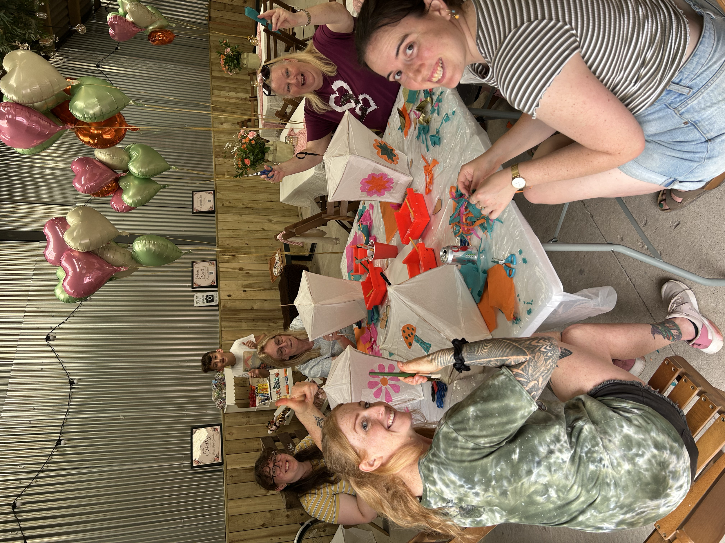 A group of smiling women and a young girl preparing paper lanterns for a celebration or event, with large pink, gold, green, and orange heart-shaped balloons floating above them, decorated tables, and floral arrangements in the background.