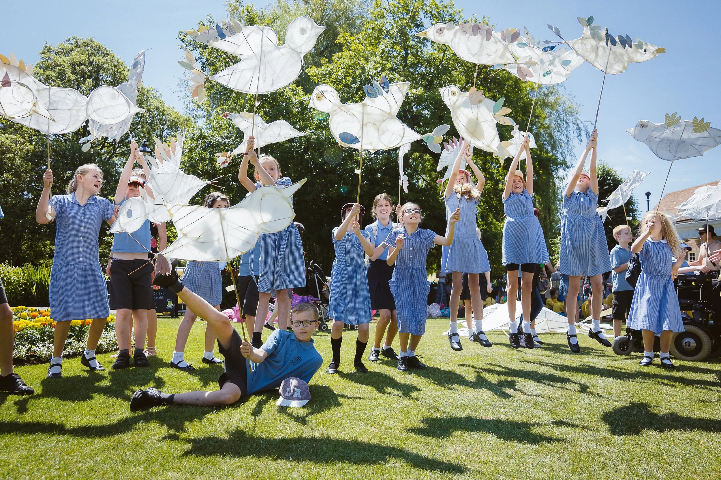 A group of children in blue school uniforms participating in a kite festival outdoors, holding large white bird-shaped kites with colorful paper accents against a background of green trees and a clear blue sky.