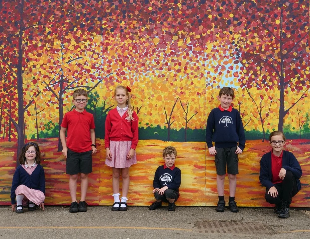 Six children in school uniforms pose in front of a colorful autumn-themed mural with orange, yellow, and red leaves and trees.