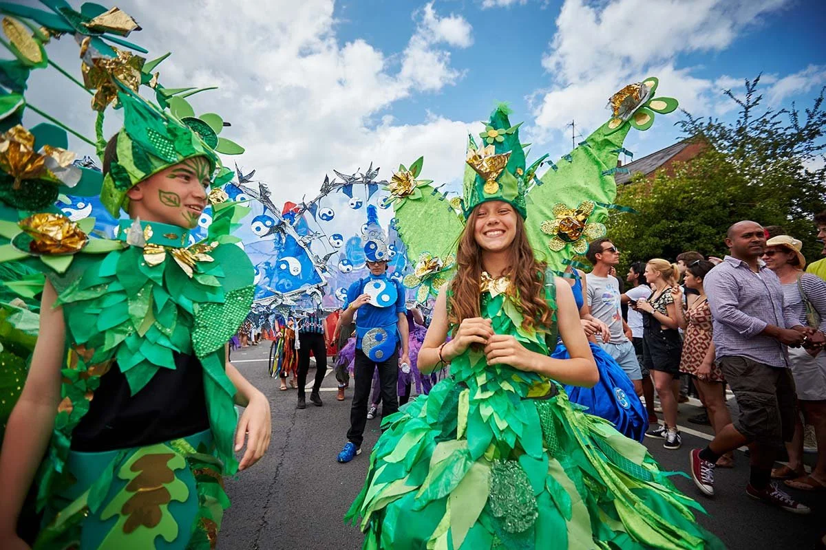 People in colorful, green, eco-friendly costumes participating in a parade or festival on a city street, with onlookers watching.