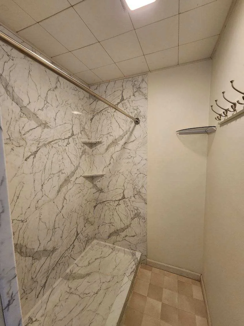 A small, empty shower with marble walls, a metal shower rod, two built-in shelves, a corner soap dish, and hook rack on the wall.