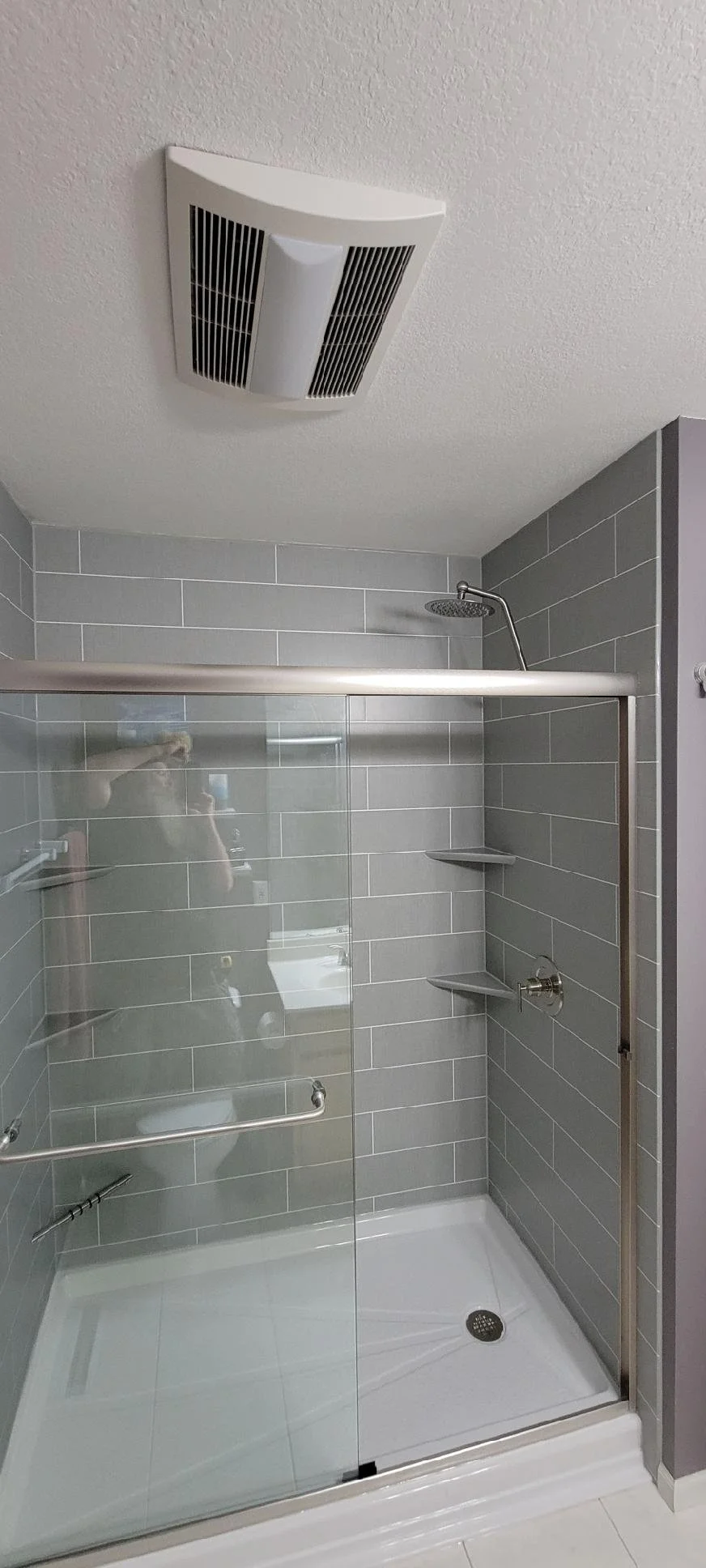 A modern shower with gray subway tile walls, a glass door, a ceiling vent, and a rain showerhead.