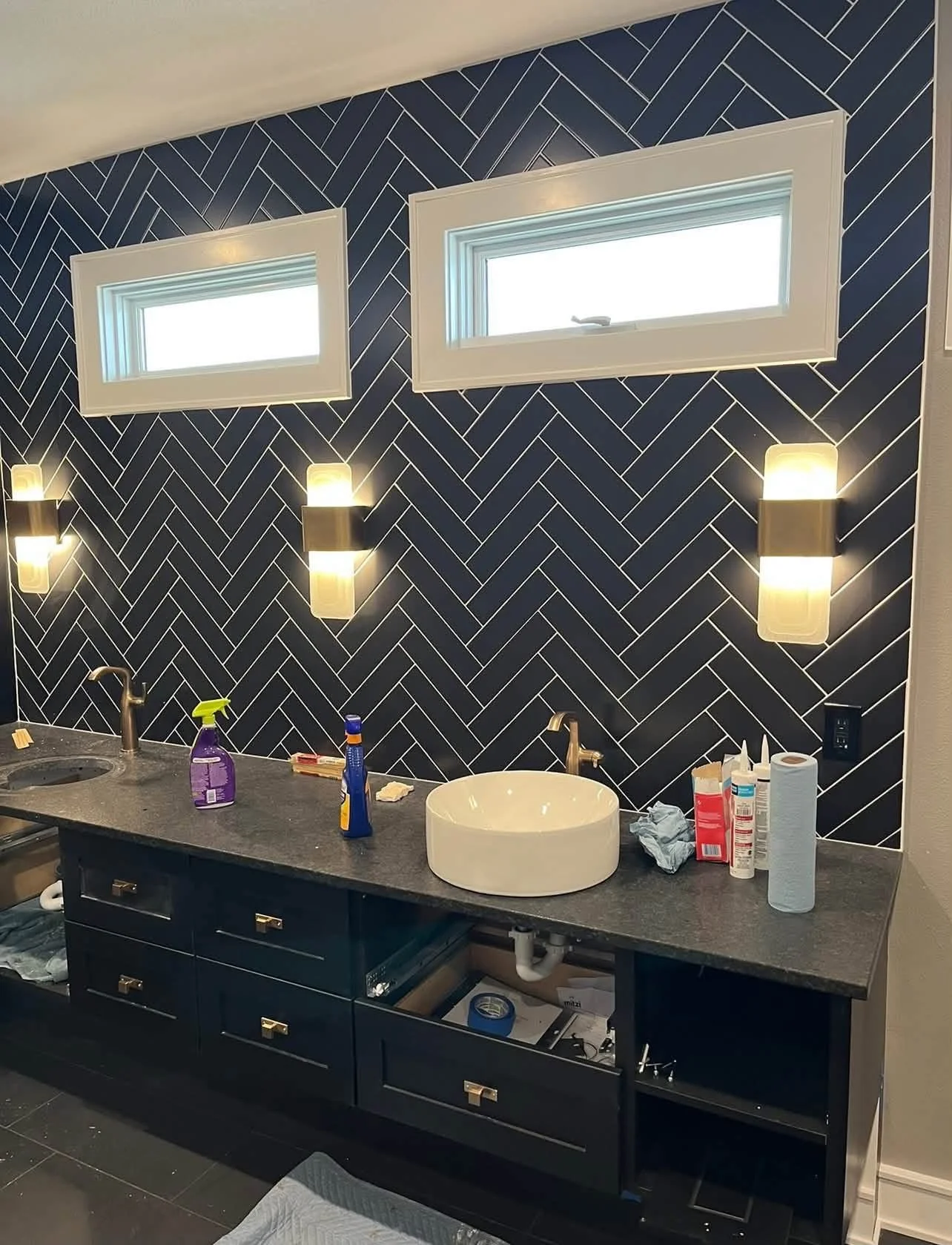Bathroom vanity area with dark blue cabinets and black countertop, two small horizontal windows, decorative herringbone patterned dark blue and white tile wall, white vessel sink, silver faucet, and two wall-mounted lights.