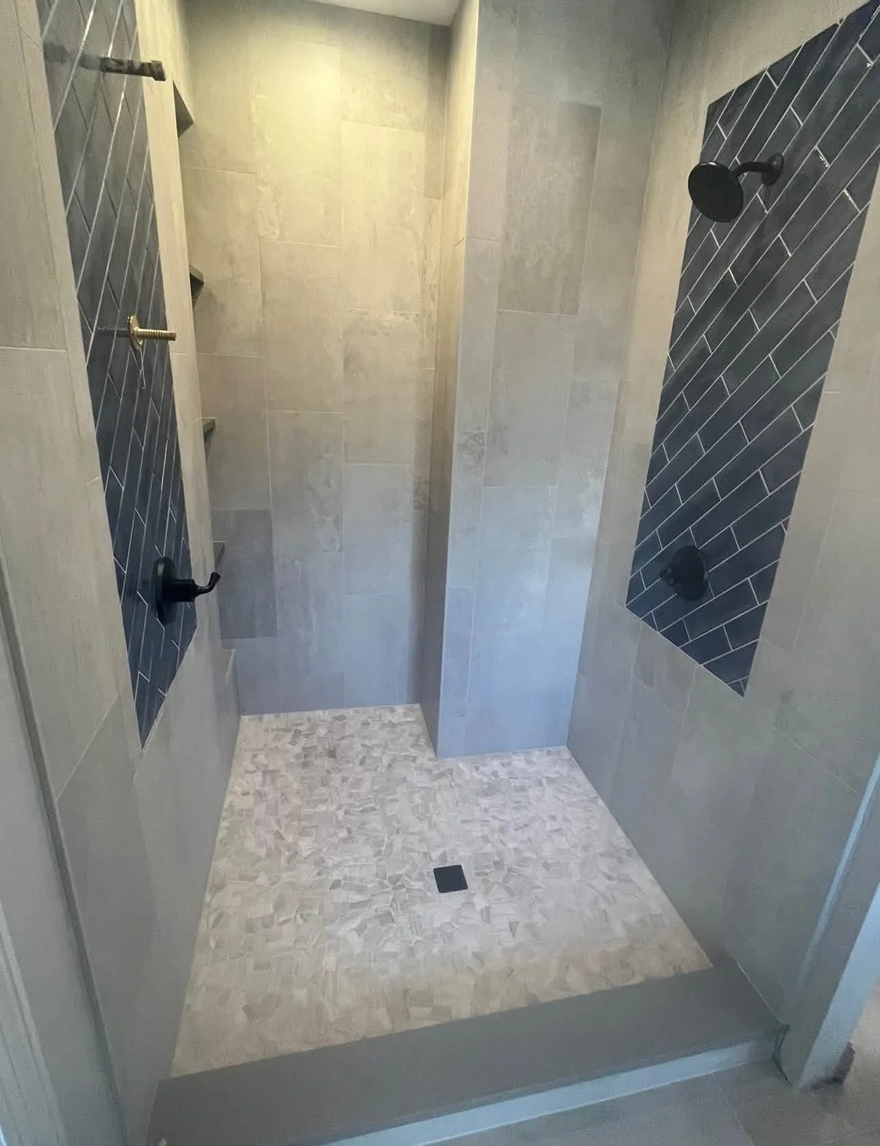 Newly renovated walk-in shower with beige and dark gray tile walls, black showerhead and controls, small mosaic tile floor, and a square floor drain.
