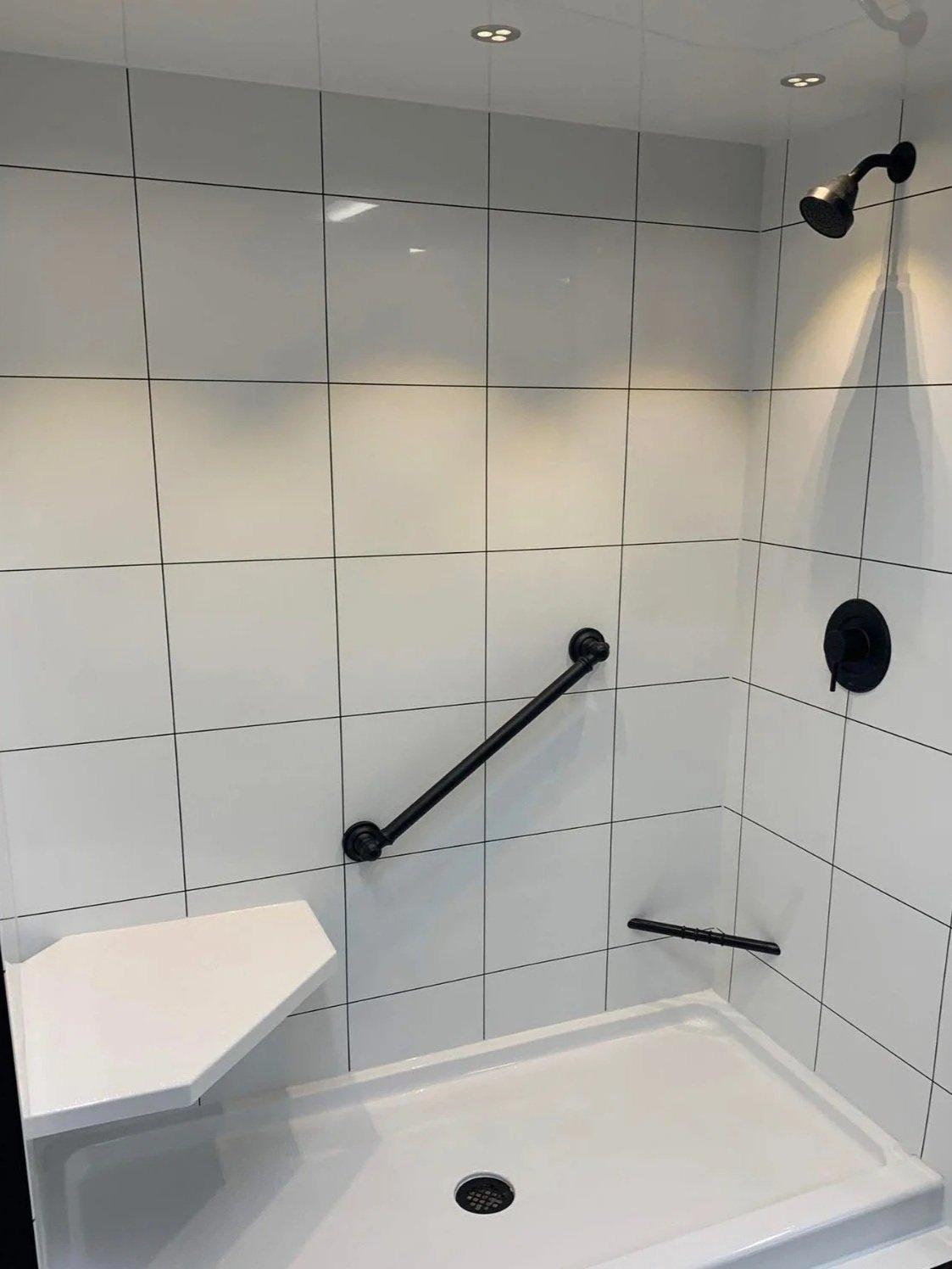 Walk-in shower with white tiled walls, black grab bar, black showerhead, black faucet, and a small white seat in the corner. A beautiful way to stay in your home and shower safely when mobility becomes more challenging. Bathroom remodel in Grimes, IA