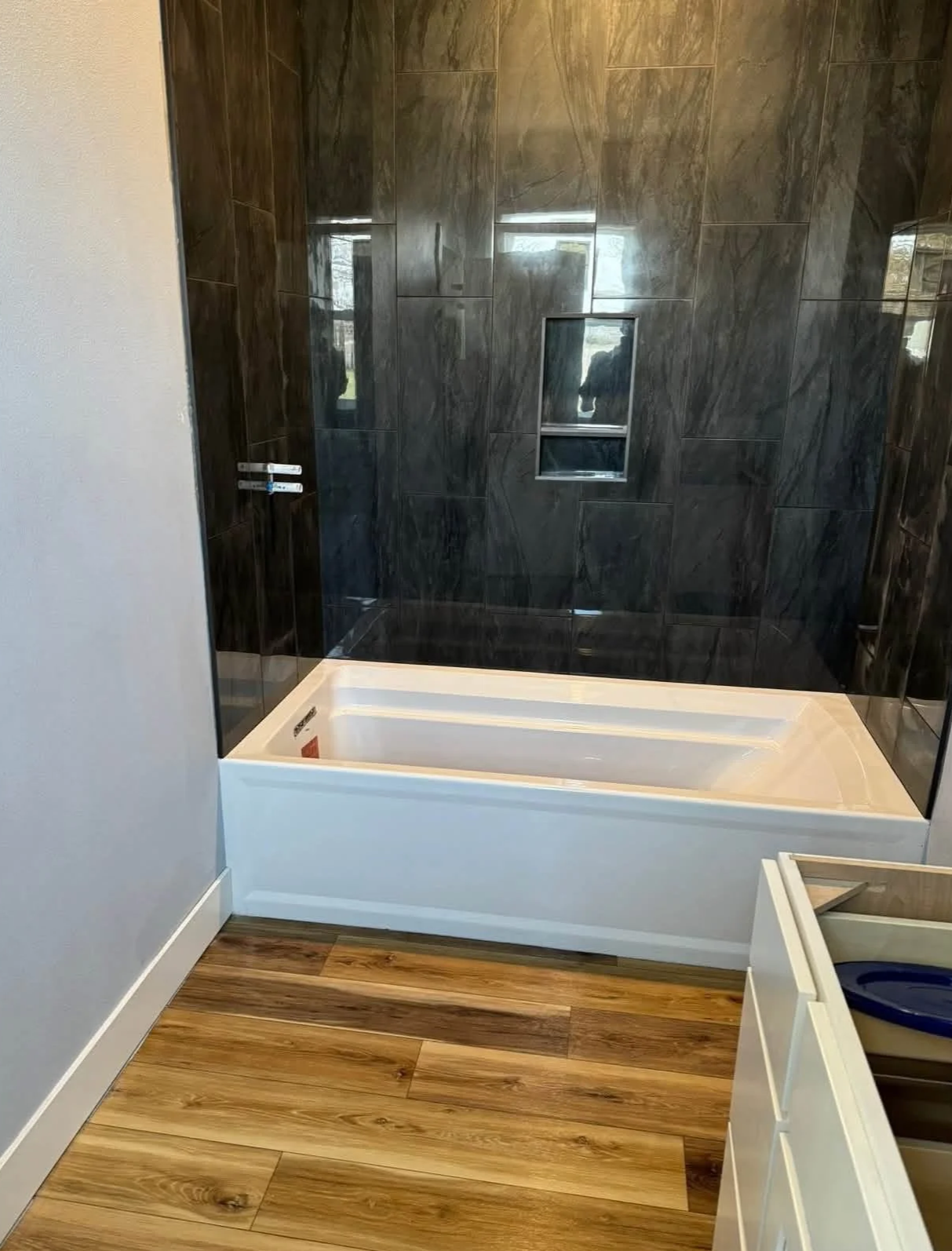 Bathroom with a partially installed bathtub and dark tiled walls. The floor has wood-like planks, and there is a small window on the wall. A dramatic bathroom update with personality in Des Moines, Iowa.