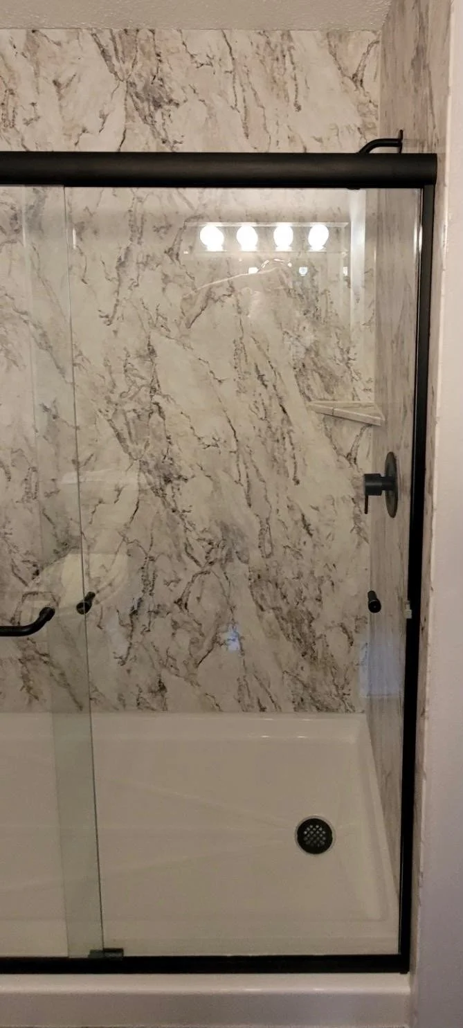 A glass shower enclosure with black hardware, featuring a marble-patterned wall and a white shower base with a drain.