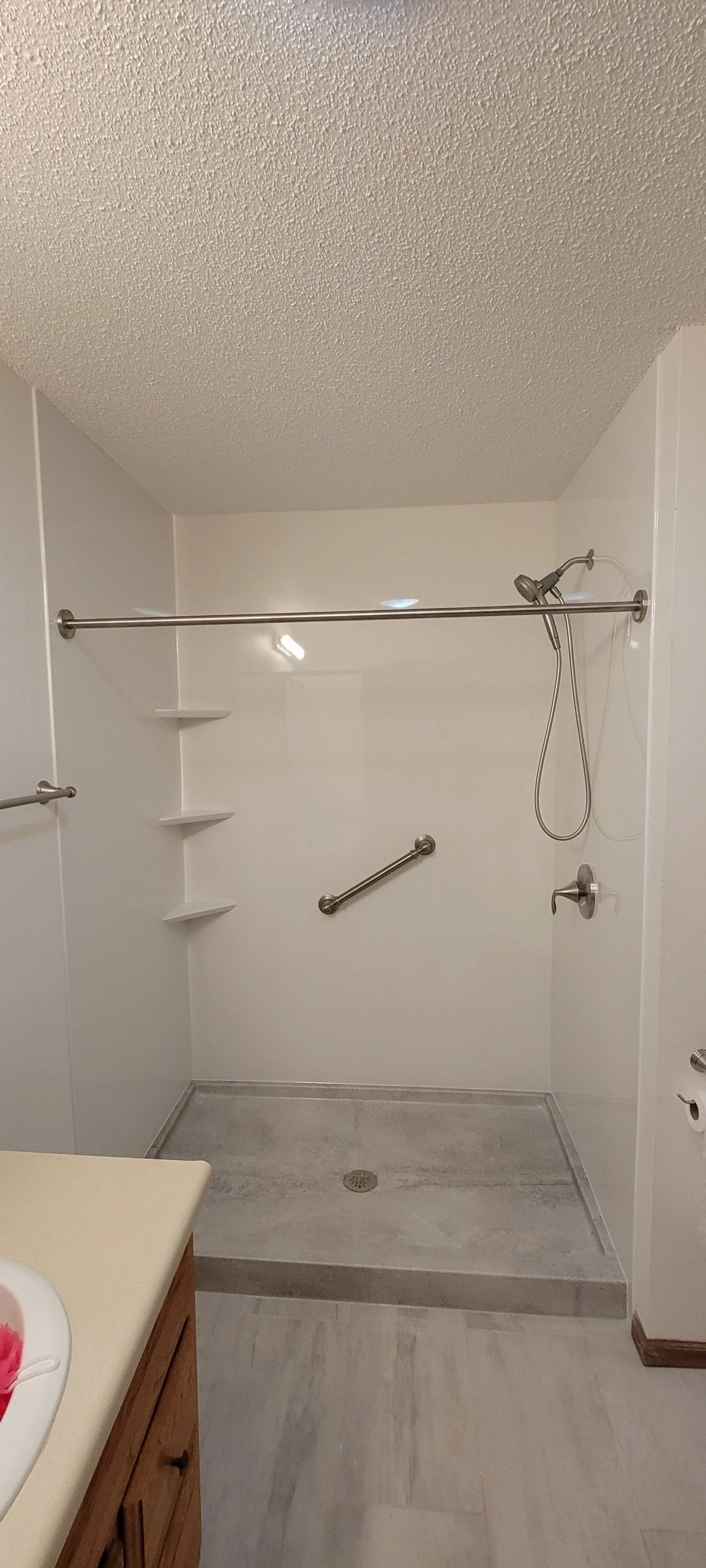 A walk-in shower with white walls, a textured ceiling, a small built-in corner shelf, handrails, a handheld shower head, and a drain in the center of the floor.
