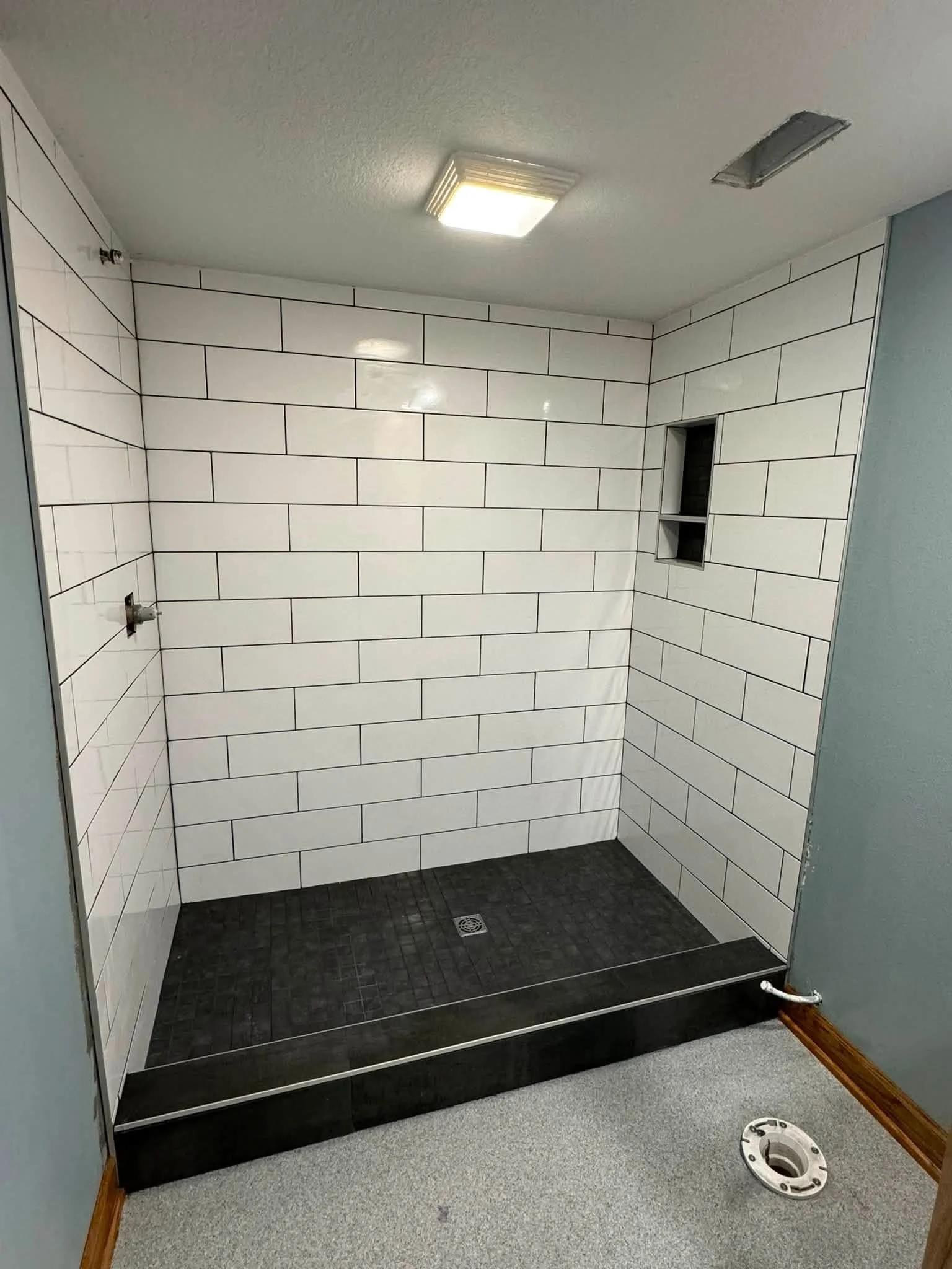 Interior of a shower stall under construction with white subway tiles on the wall, a black tiled floor, and an opening for a shower fixture. The ceiling has a bright ceiling light and an empty space for a vent, with a drain in the floor and a white PVC pipe at the bottom right.