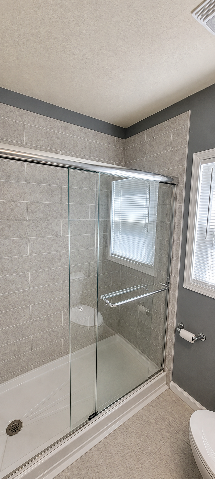 Bathroom with a glass shower enclosure, gray tiled walls, a window with blinds, and a toilet on the right.
