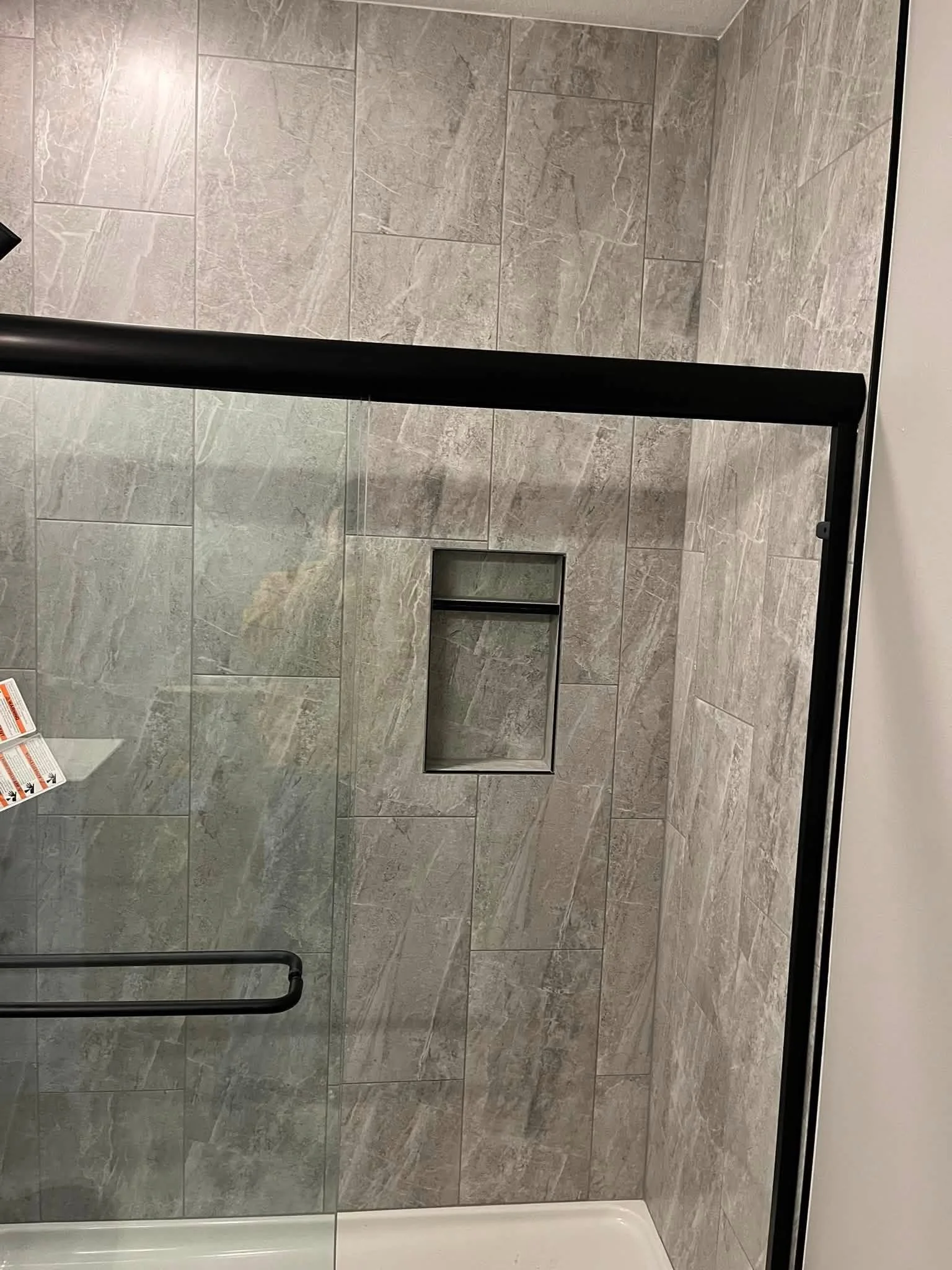A bathroom shower with gray marble tiles, a black metal shower handle, and a glass door with a black frame. There is a small built-in shelf in the tiled wall and a white shower base.