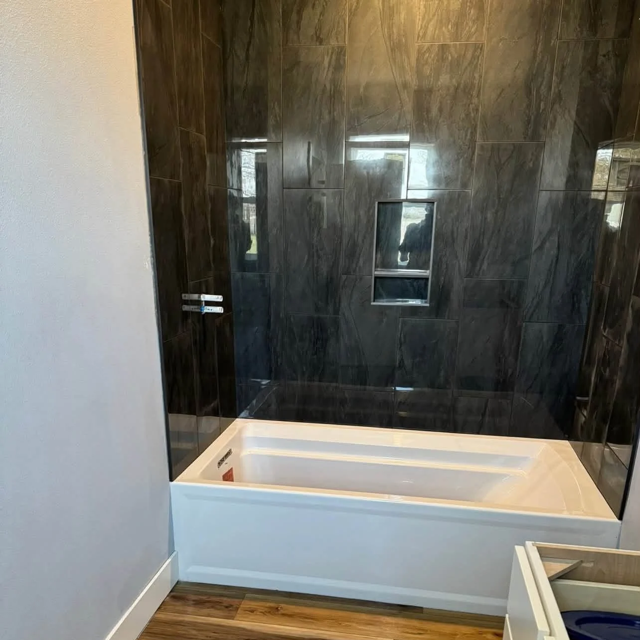 Bathroom with black marble walls, window, and a white bathtub, with a wood floor.