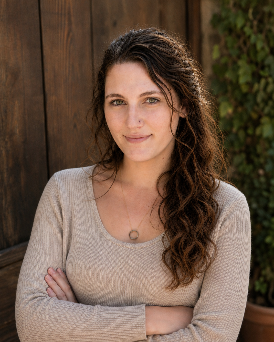 A woman with long, wavy brown hair and light green eyes standing outdoors with arms crossed, in front of a wooden fence and some greenery, wearing a beige long-sleeved top and a round necklace.