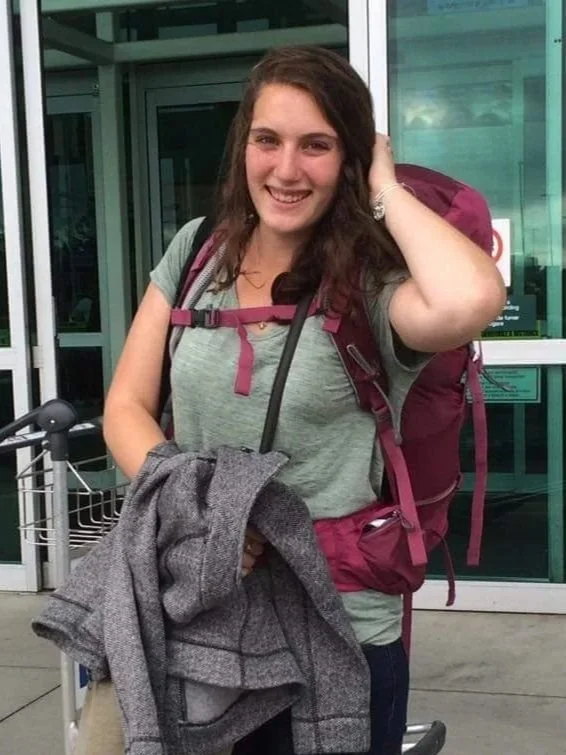 Young woman standing outside an airport terminal with a large maroon backpack and holding a gray jacket.