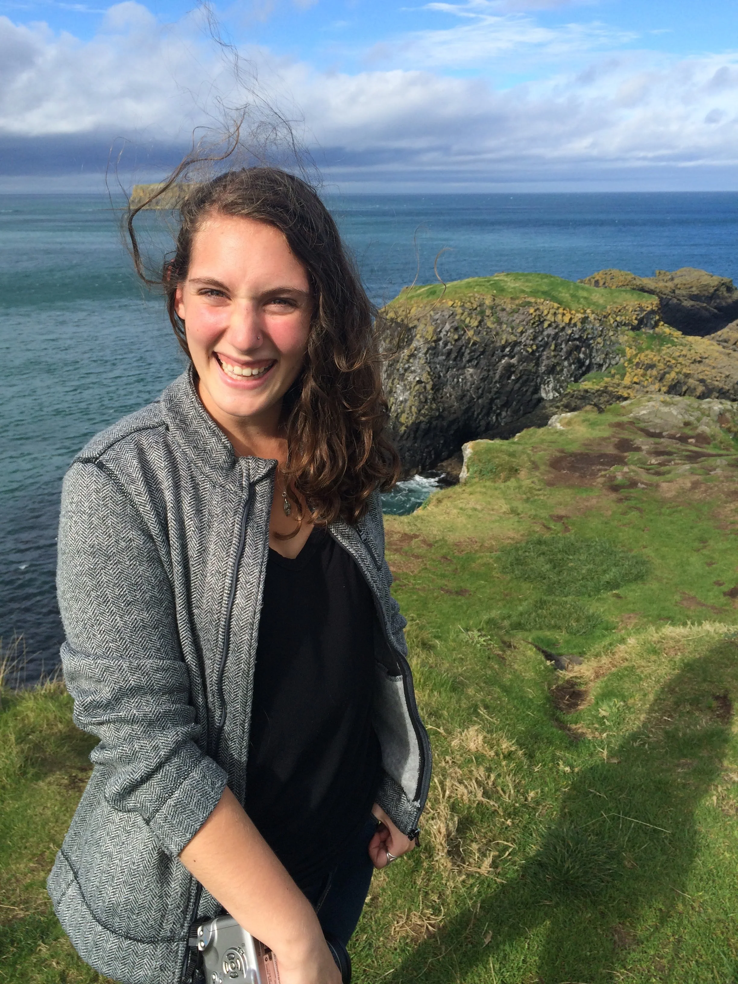 A young woman with long, curly hair smiling on a grassy cliffside by the ocean, with rocks and waves in the background under cloudy skies.