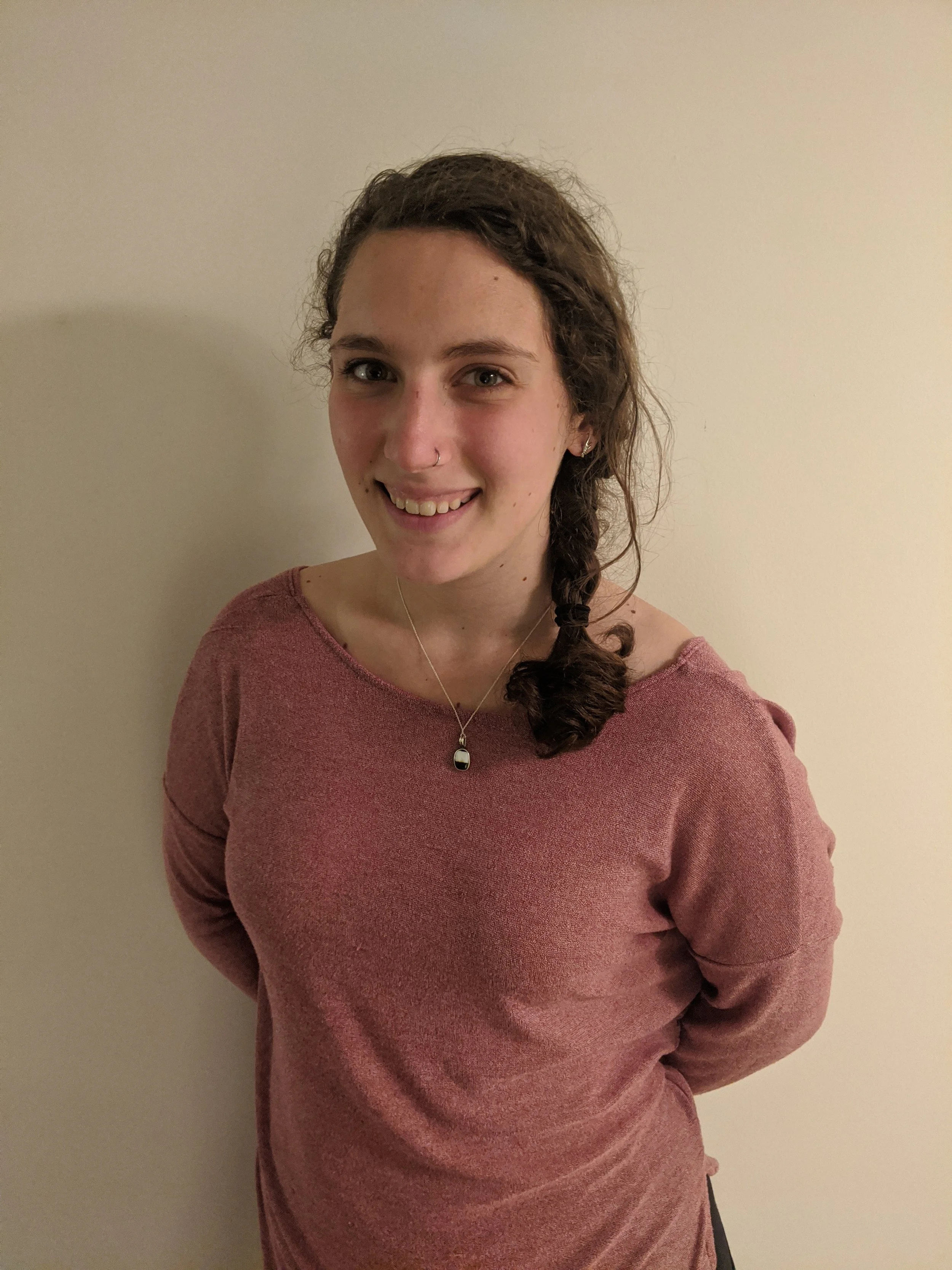 A young woman with curly brown hair in a side braid, wearing a pink off-the-shoulder sweater, silver necklace, and earrings, smiling at the camera against a plain off-white wall.