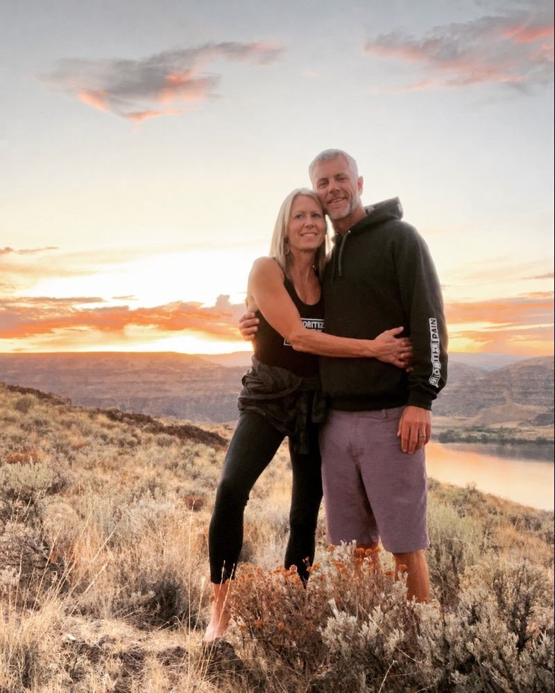 A middle-aged couple standing together outdoors during sunset, embracing each other, with a landscape of hills and a river in the background.