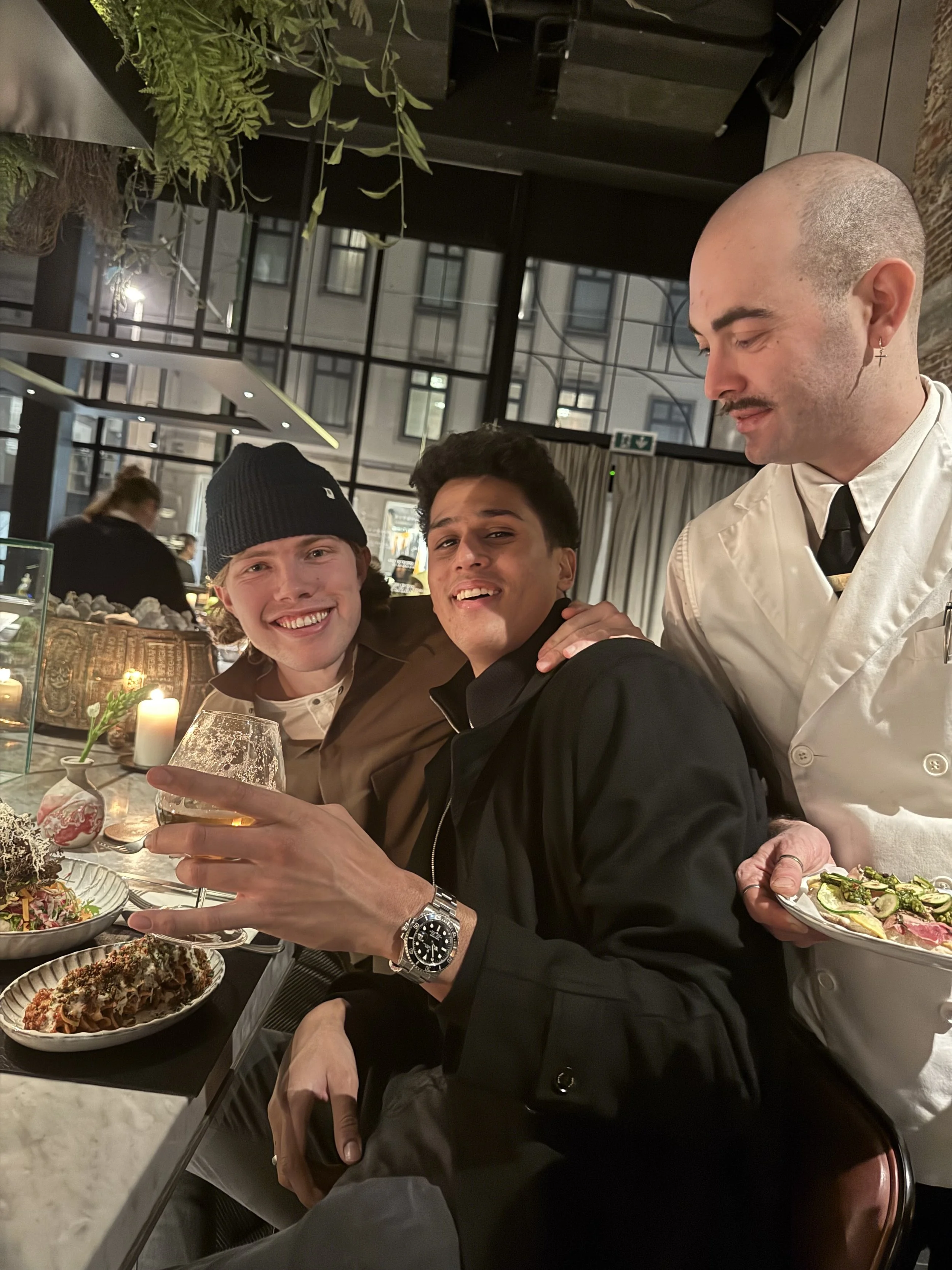 Three men are sitting at a restaurant table, smiling and enjoying food and drinks. One man is holding a glass of beer, and there are various dishes on the table, including salads and desserts. The waiter in a white uniform is standing next to the middle man, smiling. The background shows a modern interior with large windows and a brick wall.