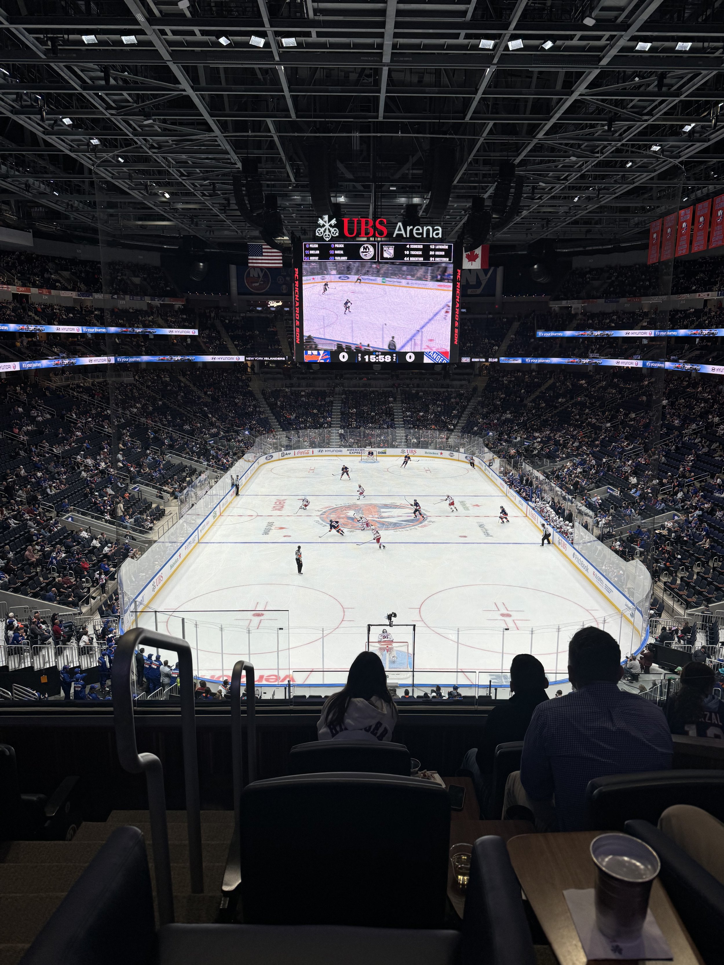 Inside UBS Arena with spectators watching a hockey game on the ice rink. The picture is taken from the seating area, showing rowdy crowds, a large scoreboard, and banners hanging from the ceiling.