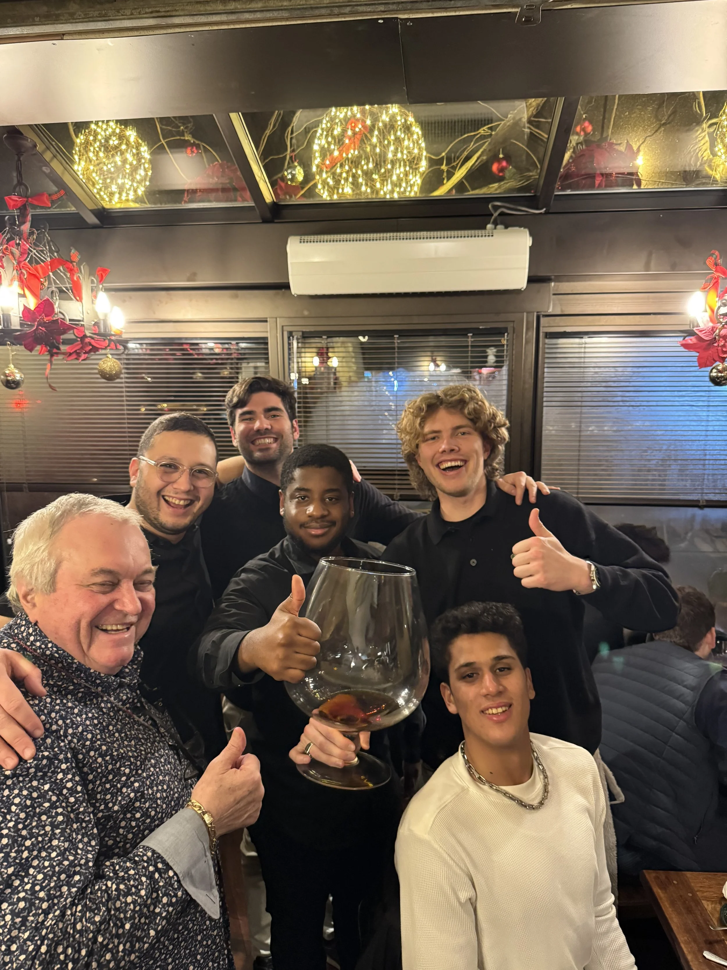A group of smiling people celebrating at a festive gathering, with some giving thumbs up, holding a large glass of wine, and surrounded by Christmas decorations and warm lighting.