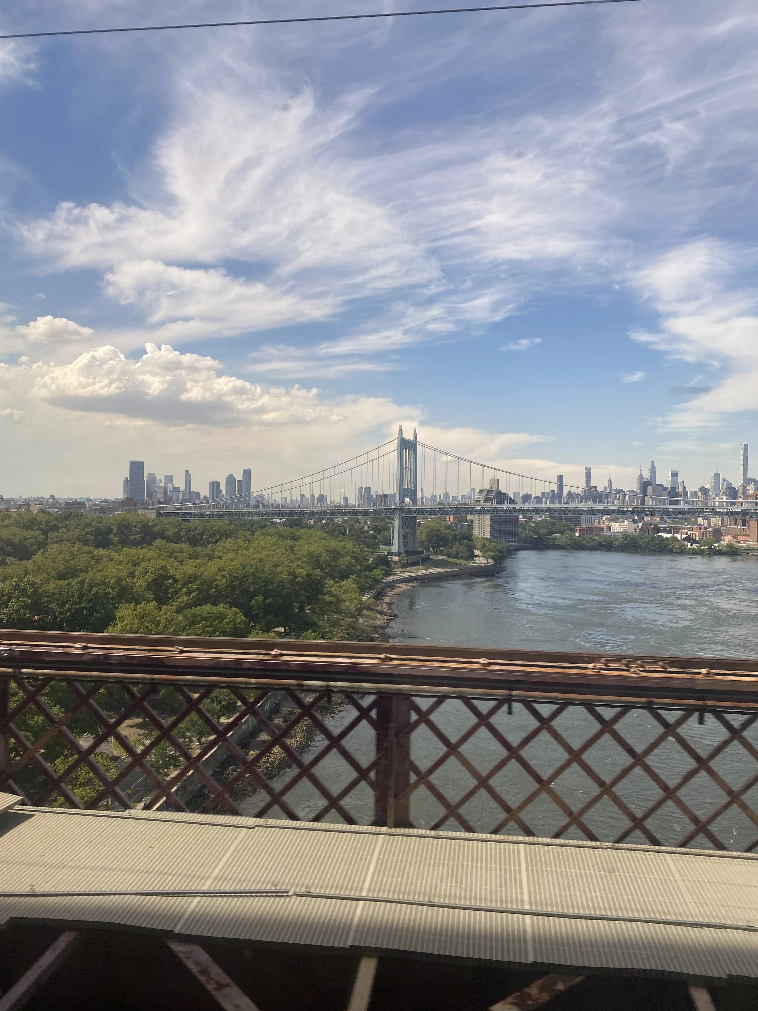 View of New York City skyline with a bridge over a river, clouds in the sky, and a section of a bridge or overpass with railings in the foreground.