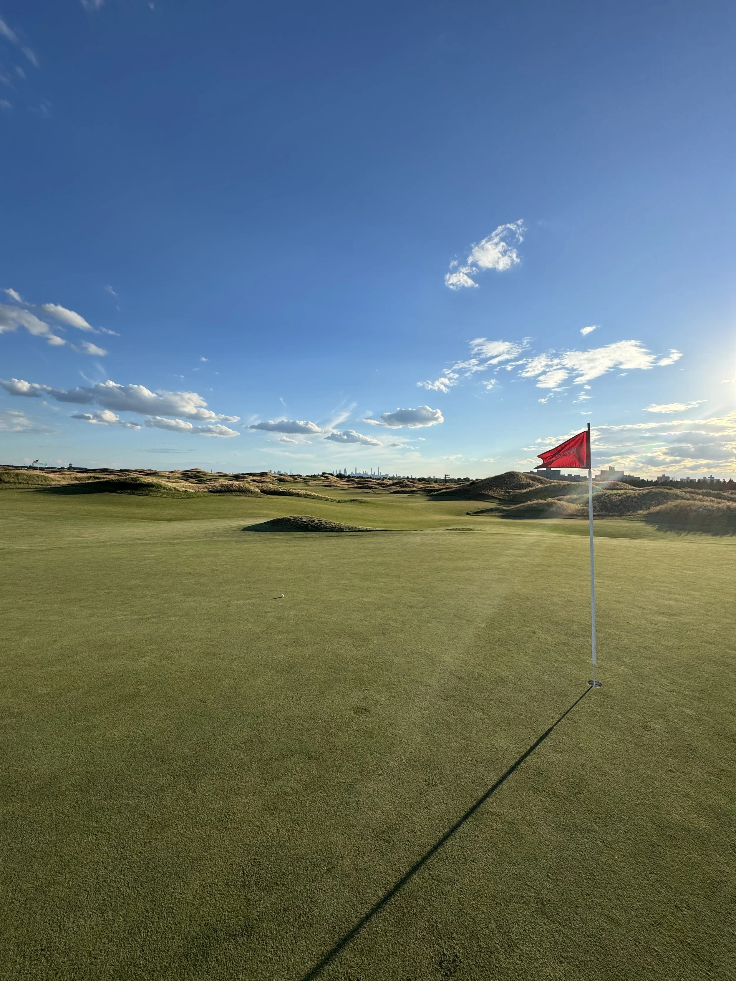 A golf course with a red flag on a green, under a partly cloudy sky during late afternoon or early evening, with the city skyline visible in the distance.