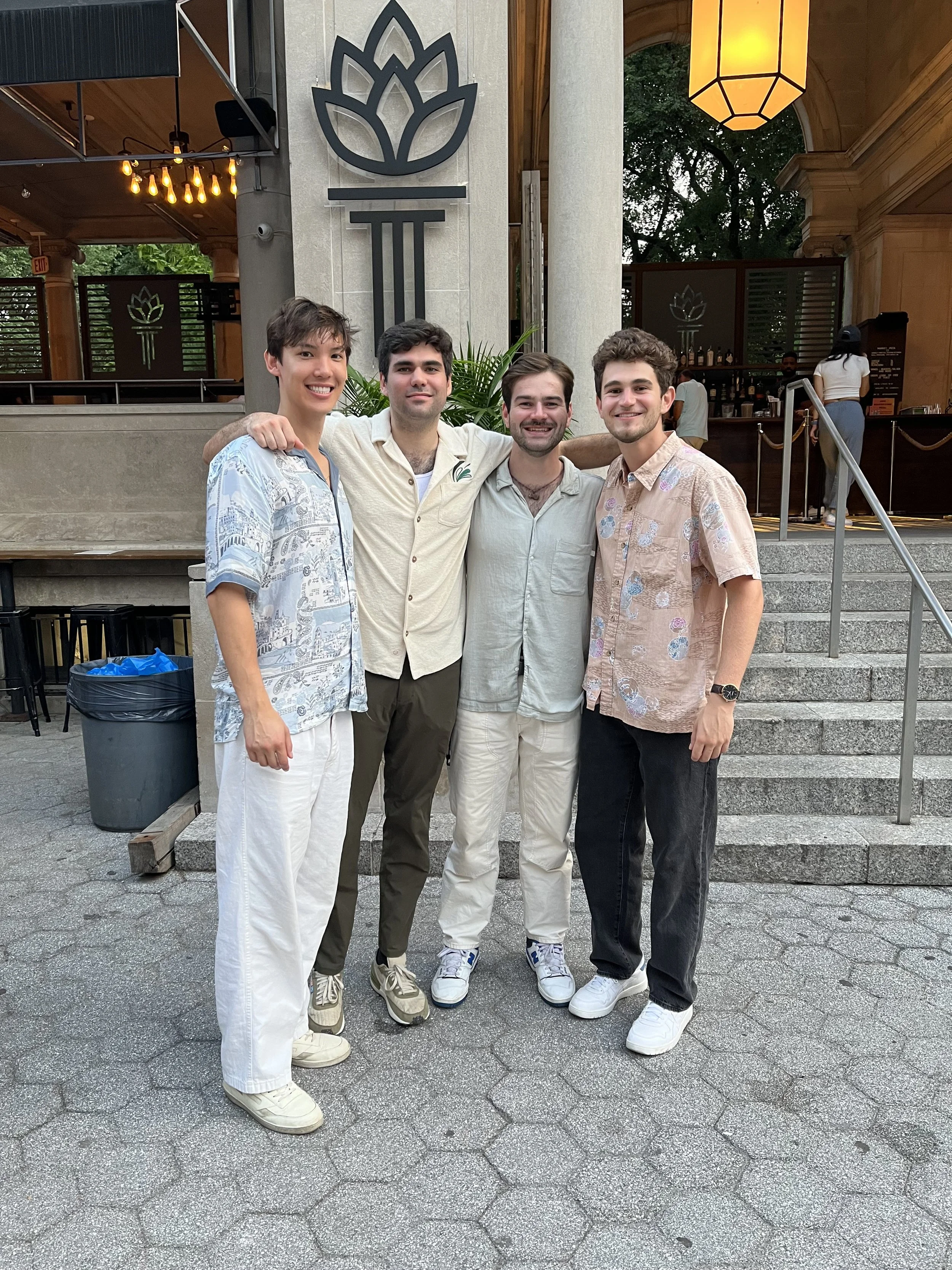 Four young men standing together outside a building, smiling at the camera. They are dressed casually and appear to be friends at a social gathering.