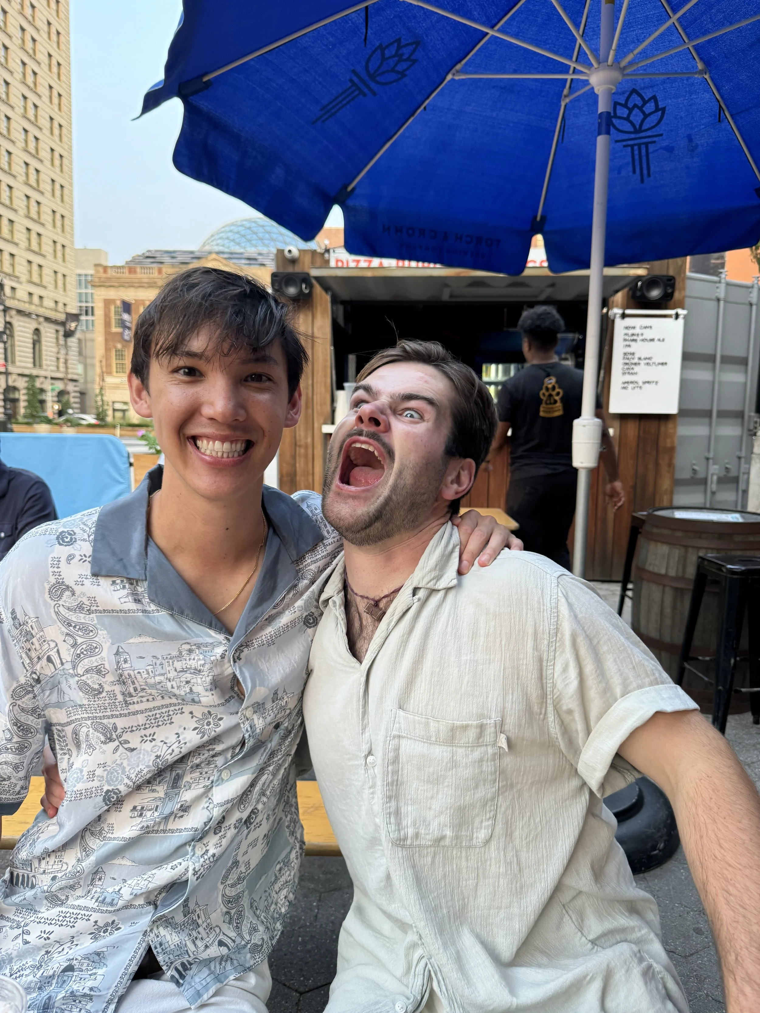 Two young men smiling and posing for a photo outdoors, with one man hugging the other, under a large blue umbrella with a cityscape background and a street scene.