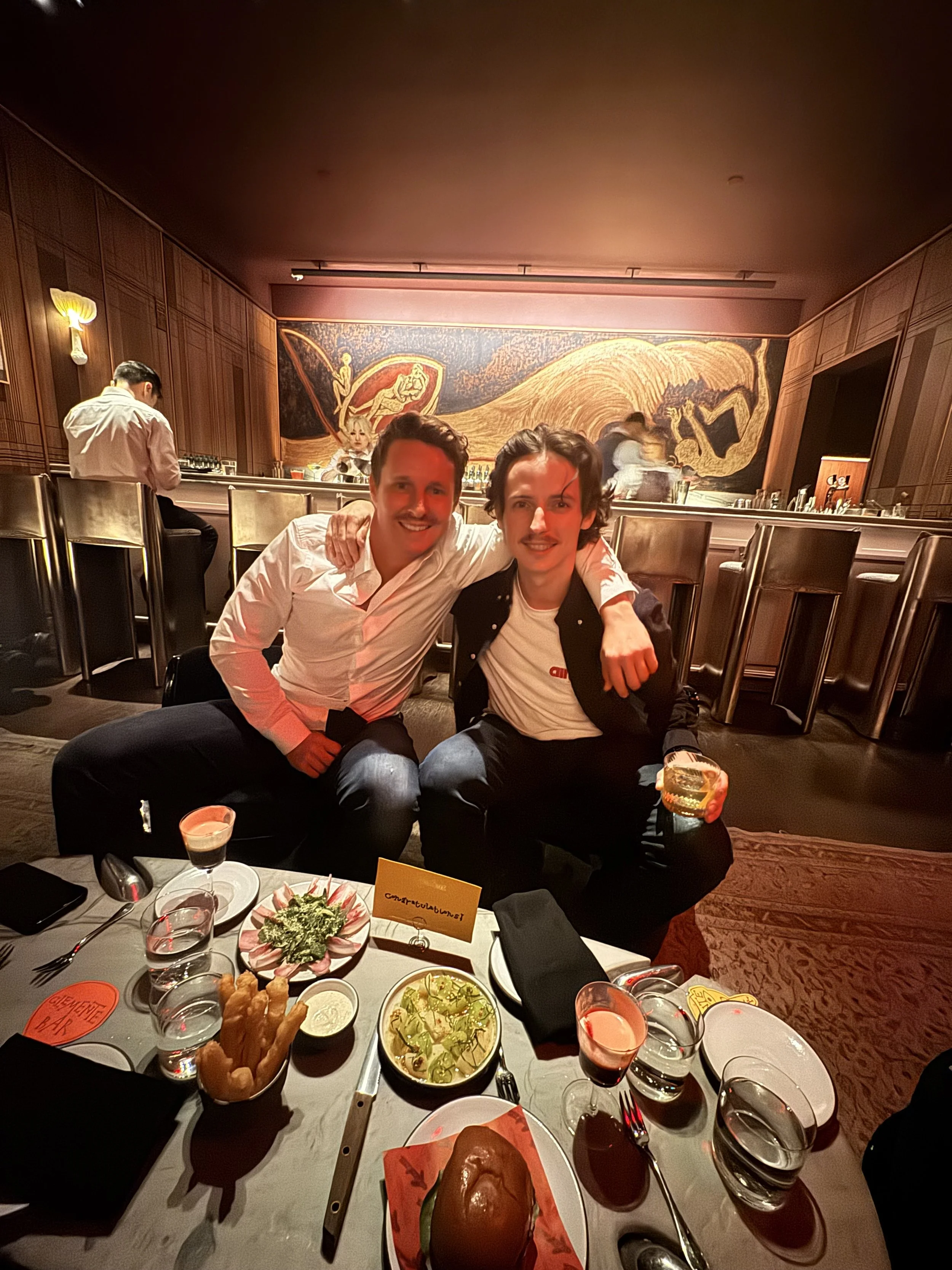 Two smiling men sitting at a dining table with various dishes, drinks, and a congratulatory note on the table. Behind them is a large mural and a bar, with a bartender attending to another customer.