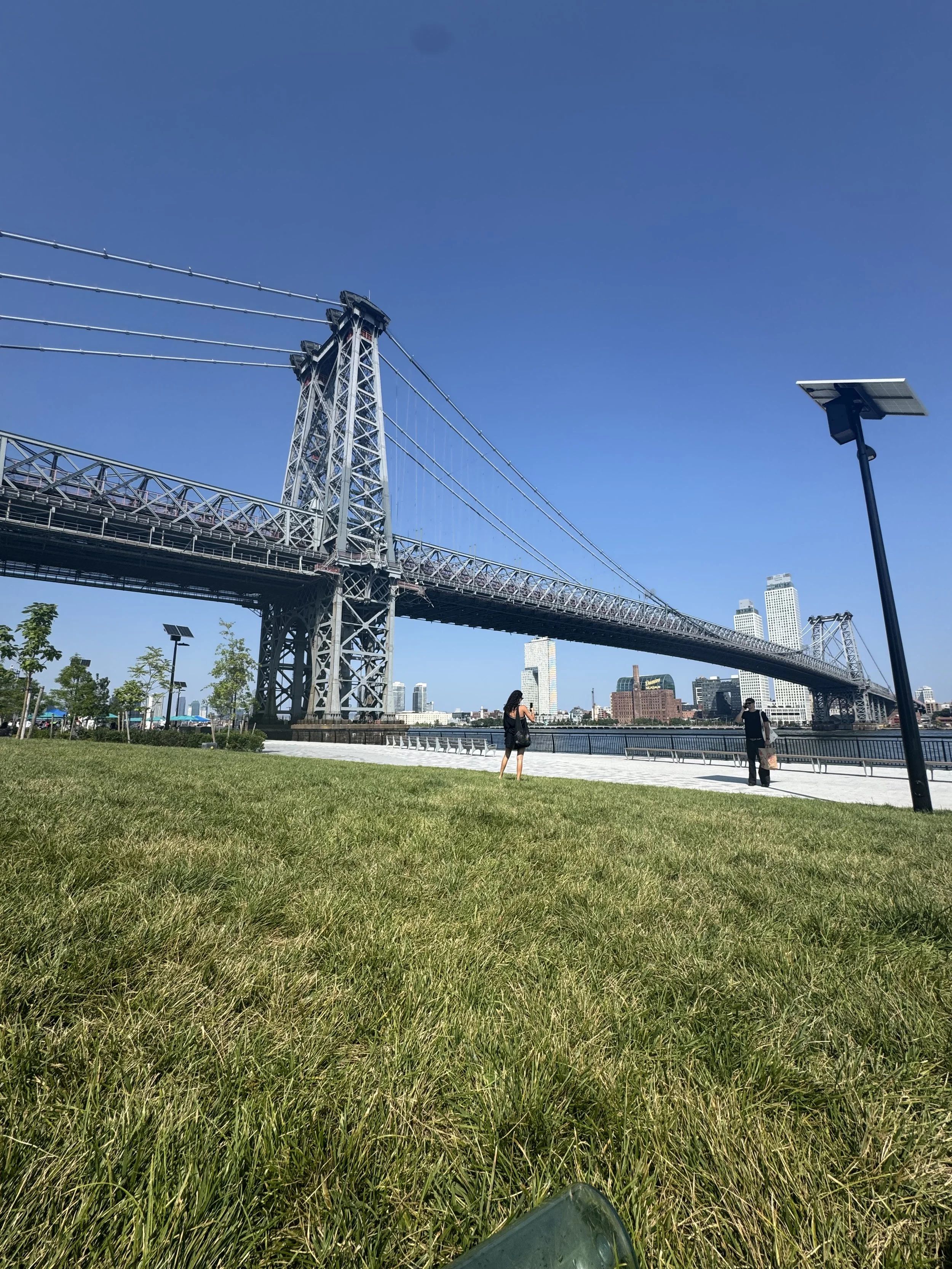Photo of the Brooklyn Bridge over the water with a city skyline in the background, taken from a park with green grass and a person standing by the water.