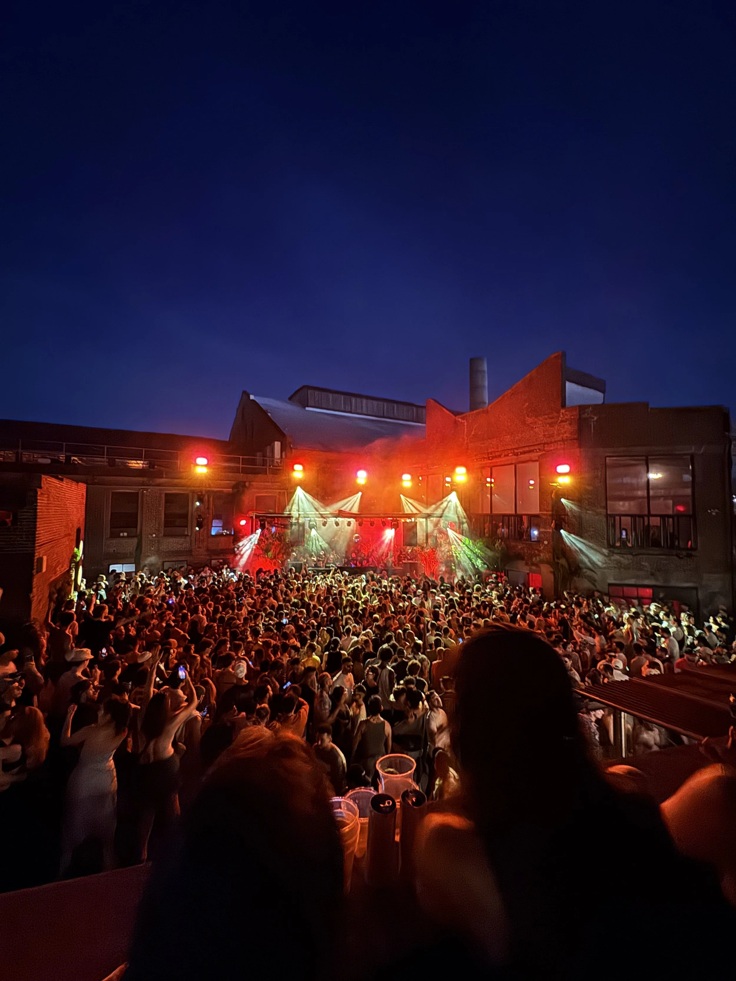 Crowd gathered at an outdoor concert stage at night, with colorful stage lights and a dark sky in the background.