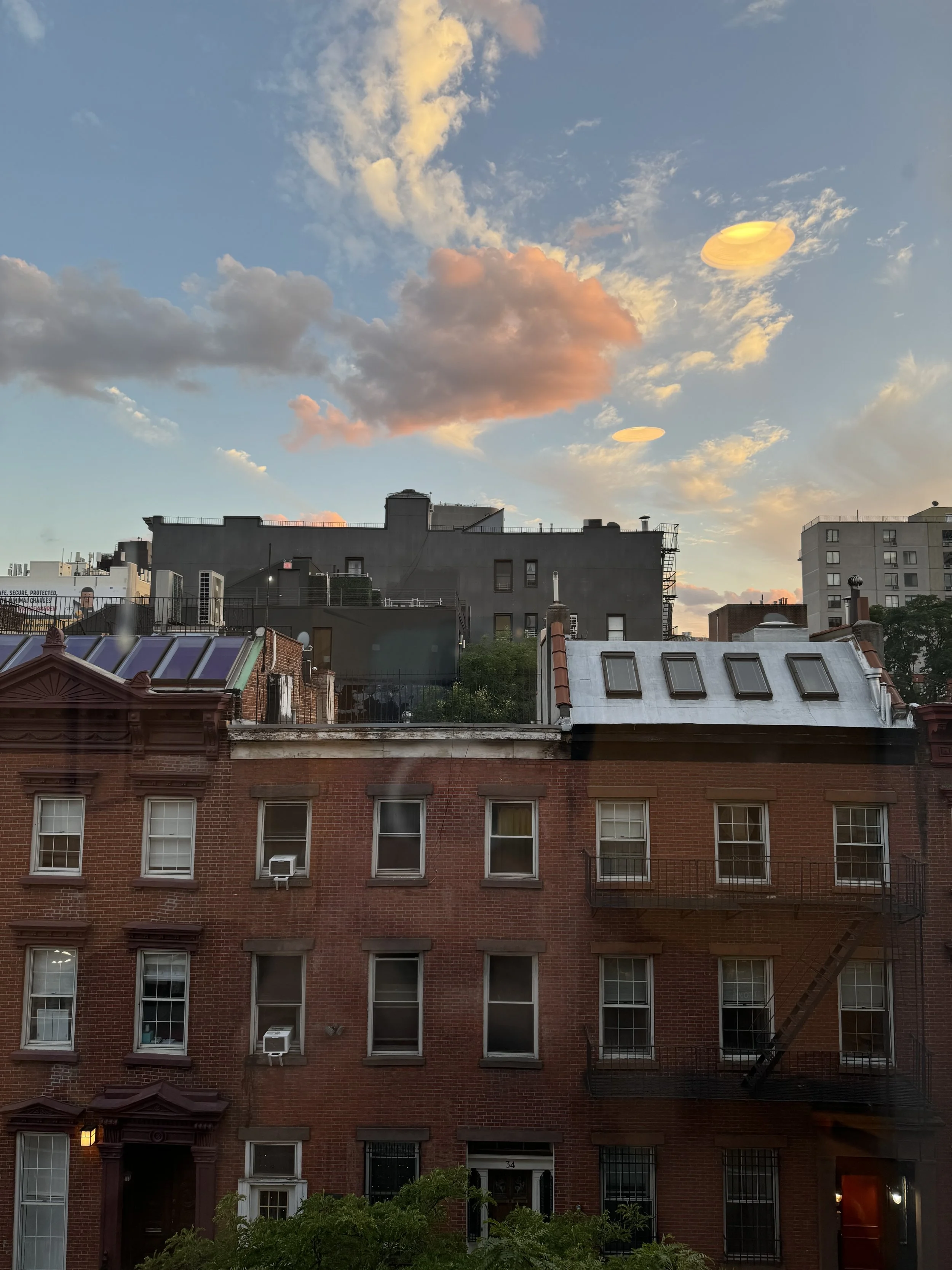 City skyline view at sunset with buildings, clouds with pink and orange hues, and reflections on the windows.