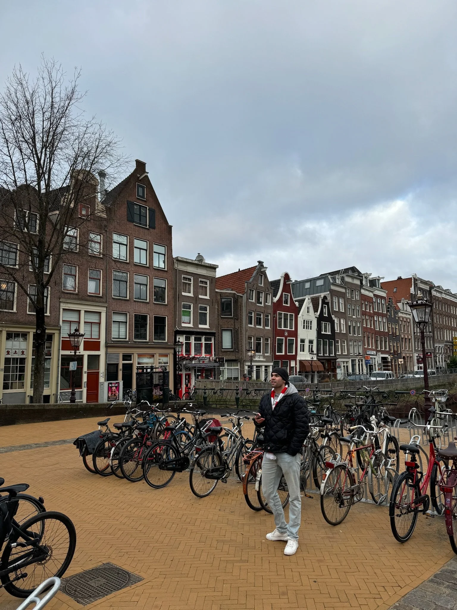 A man standing among bicycles in front of colorful traditional European townhouses with gabled roofs and large windows, under a cloudy sky.