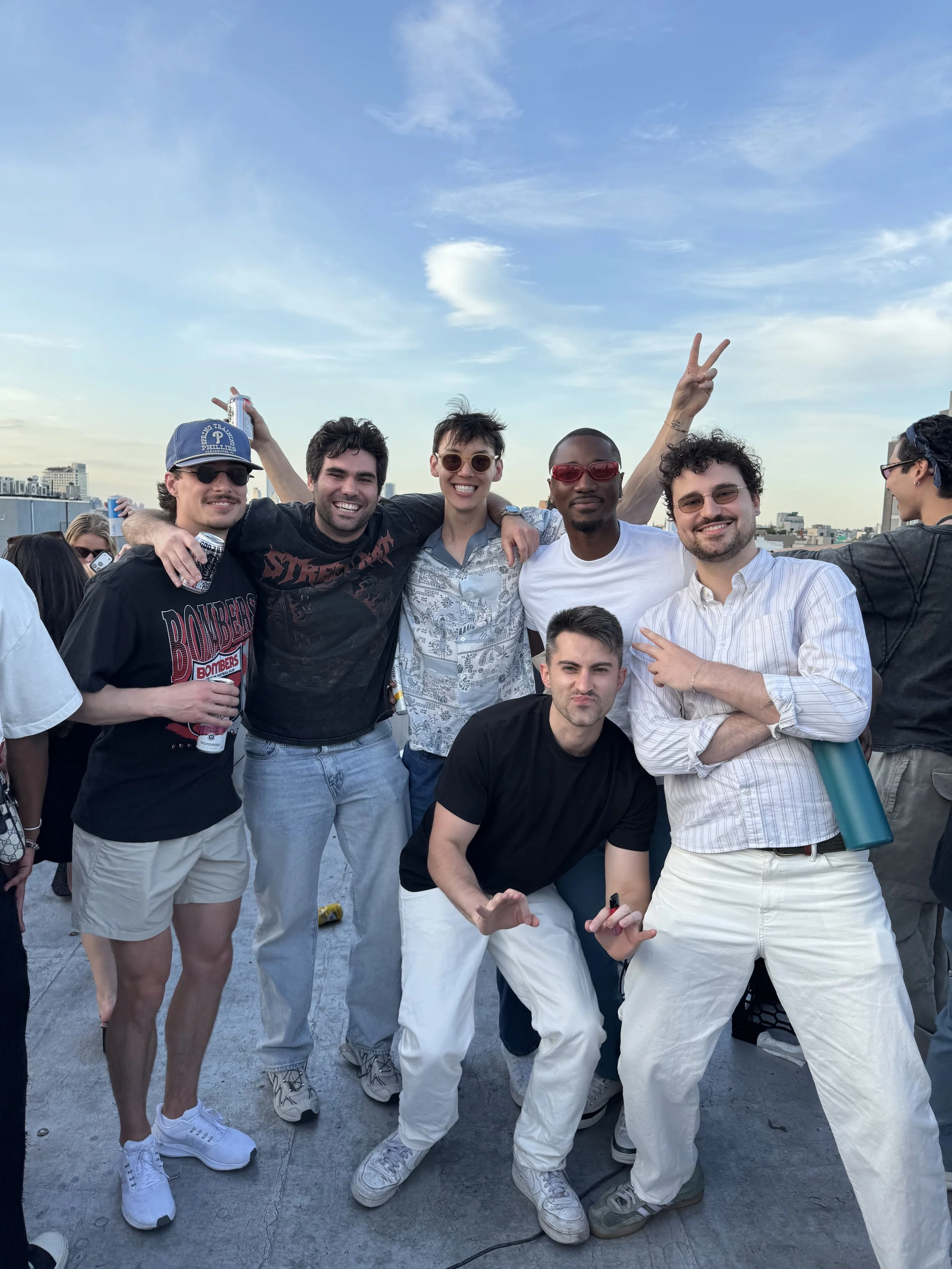 A group of friends enjoying an outdoor gathering on a rooftop or terrace with a cityscape in the background, smiling and posing for a photo.