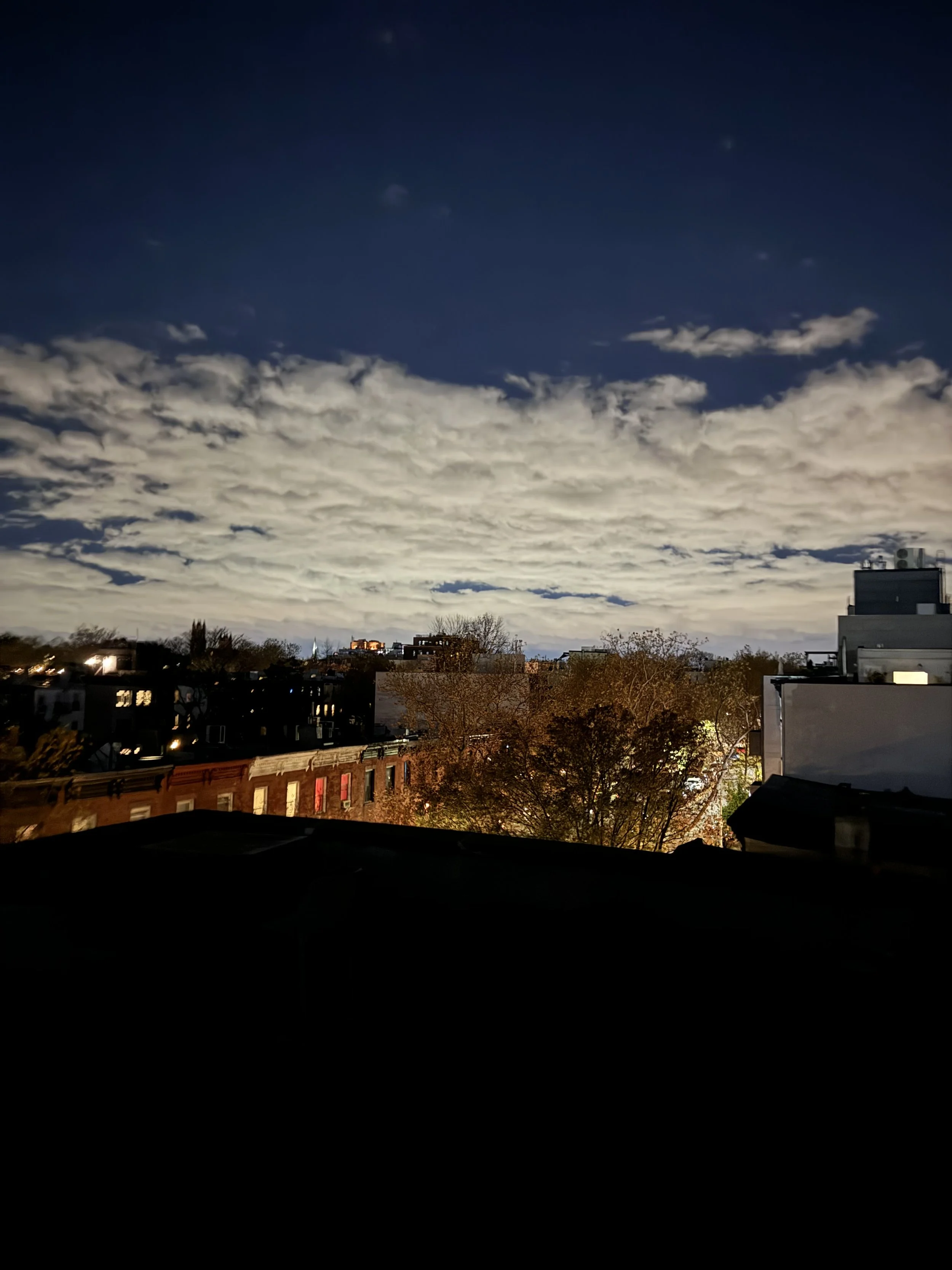 Nighttime cityscape with clouds over buildings and trees, with some illuminated windows and a dark sky.