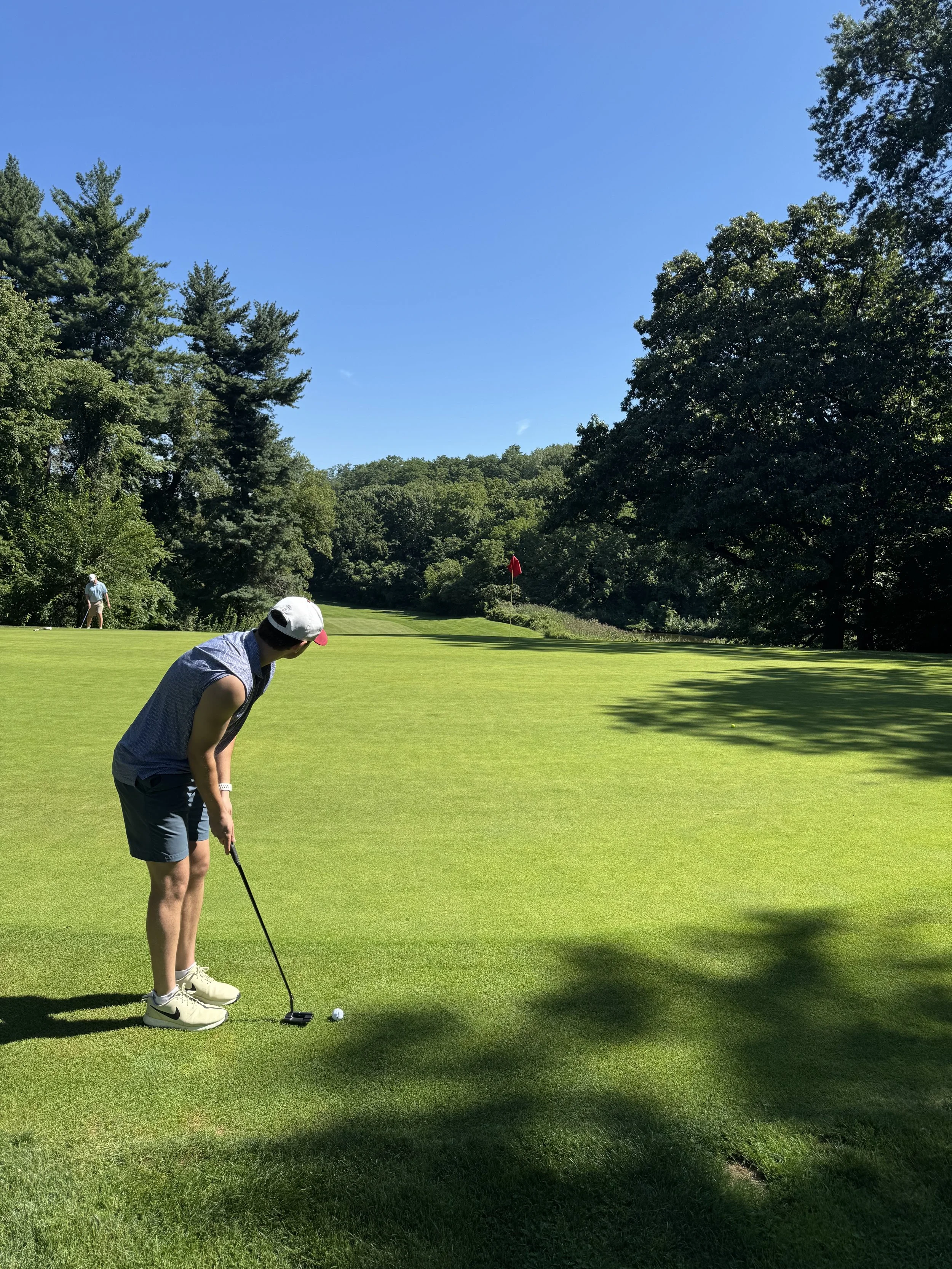 A man in a sleeveless shirt and shorts preparing to putt on a golf green, with a red flag in the background, surrounded by trees and a clear blue sky.