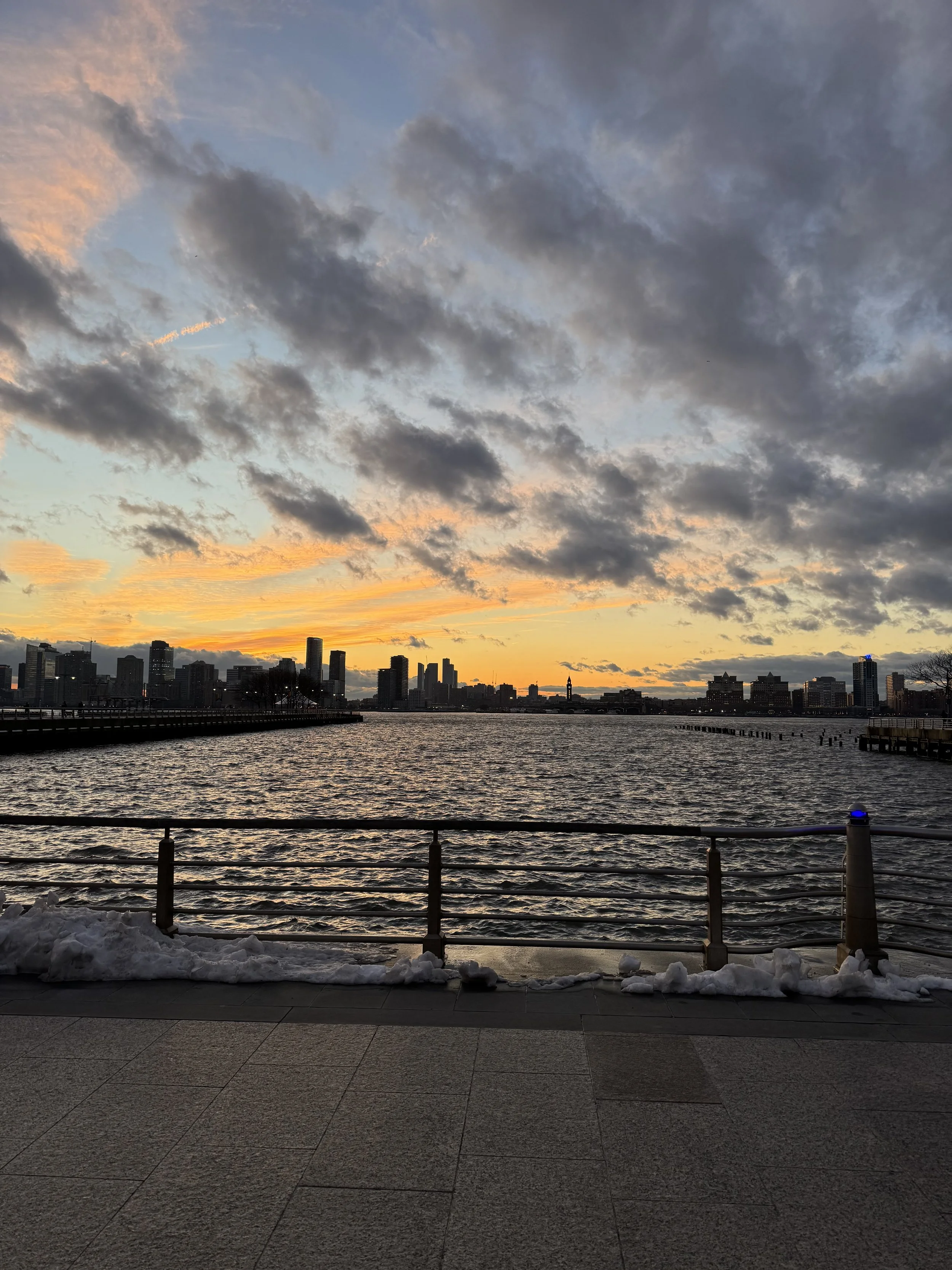 City skyline viewed from a waterfront promenade during sunset, with dark clouds in the sky and snow on the ground.