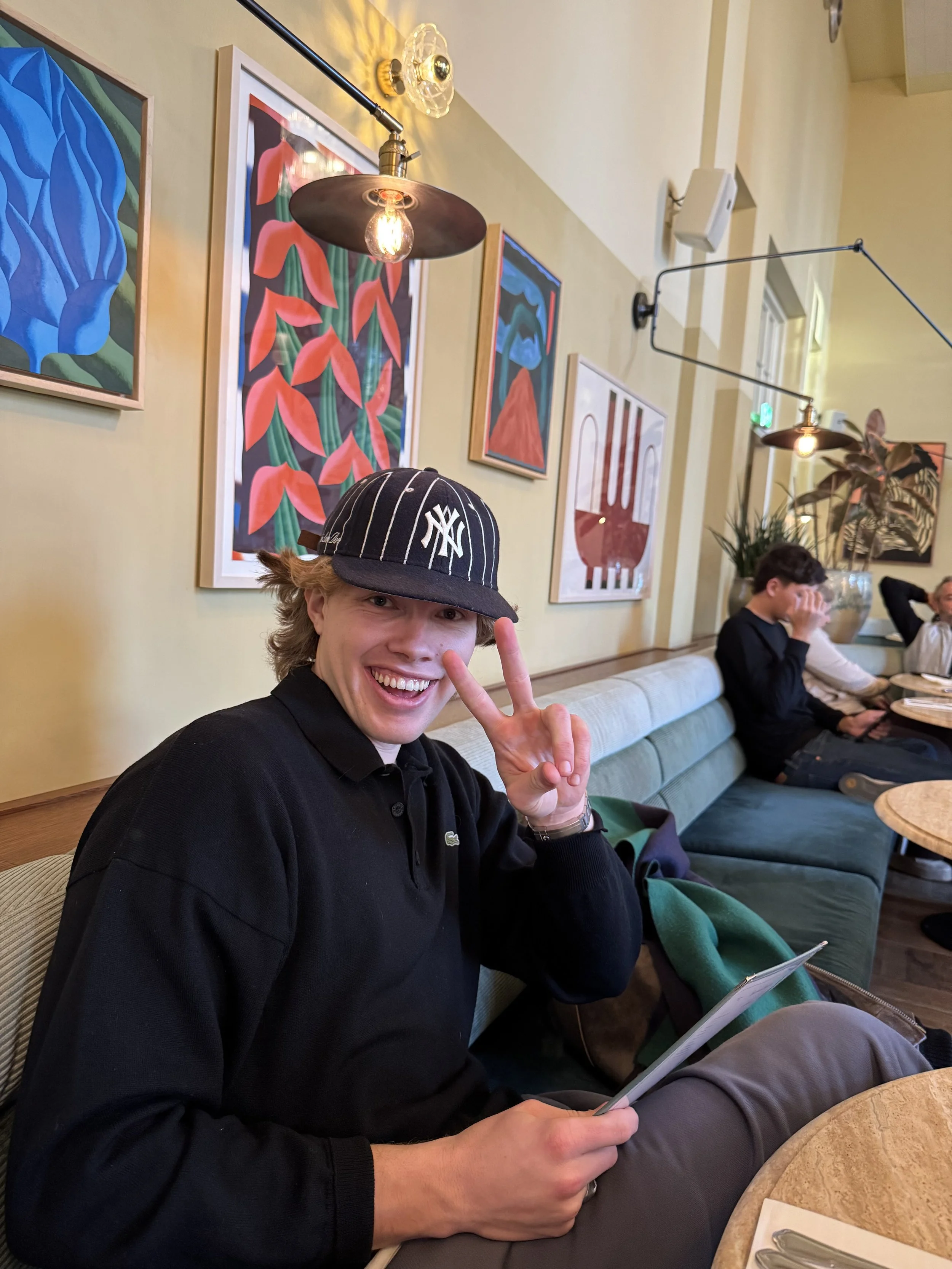 A young man smiling and making a peace sign at the camera while sitting in a restaurant or cafe with artwork on the wall behind him. He is wearing a black baseball cap and a black sweater.