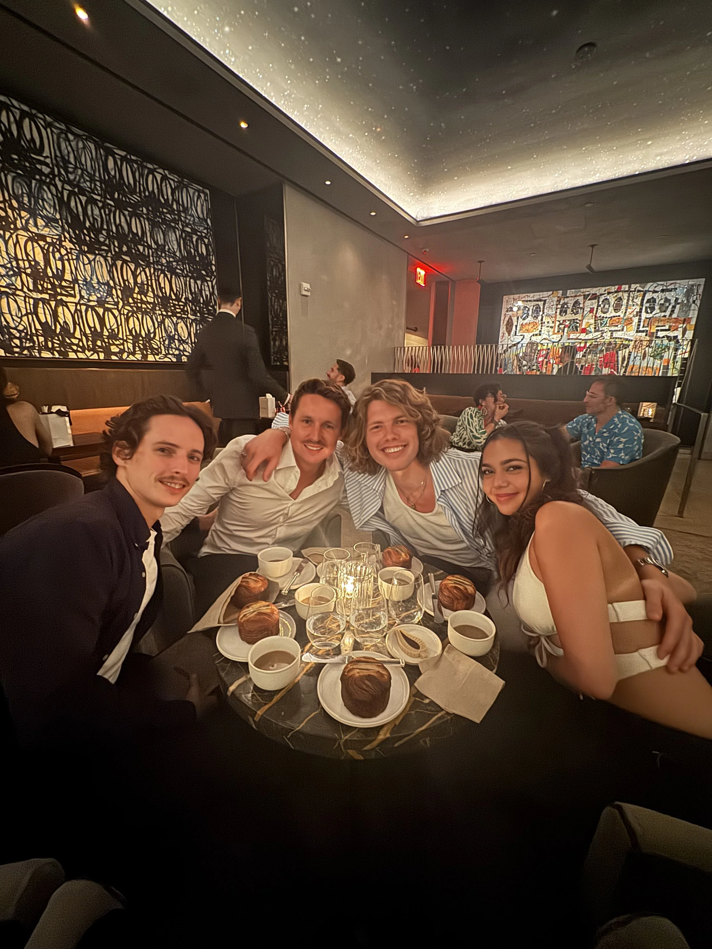 Group of five smiling young people sitting at a round table in a restaurant, with desserts and coffee cups, celebrating together.
