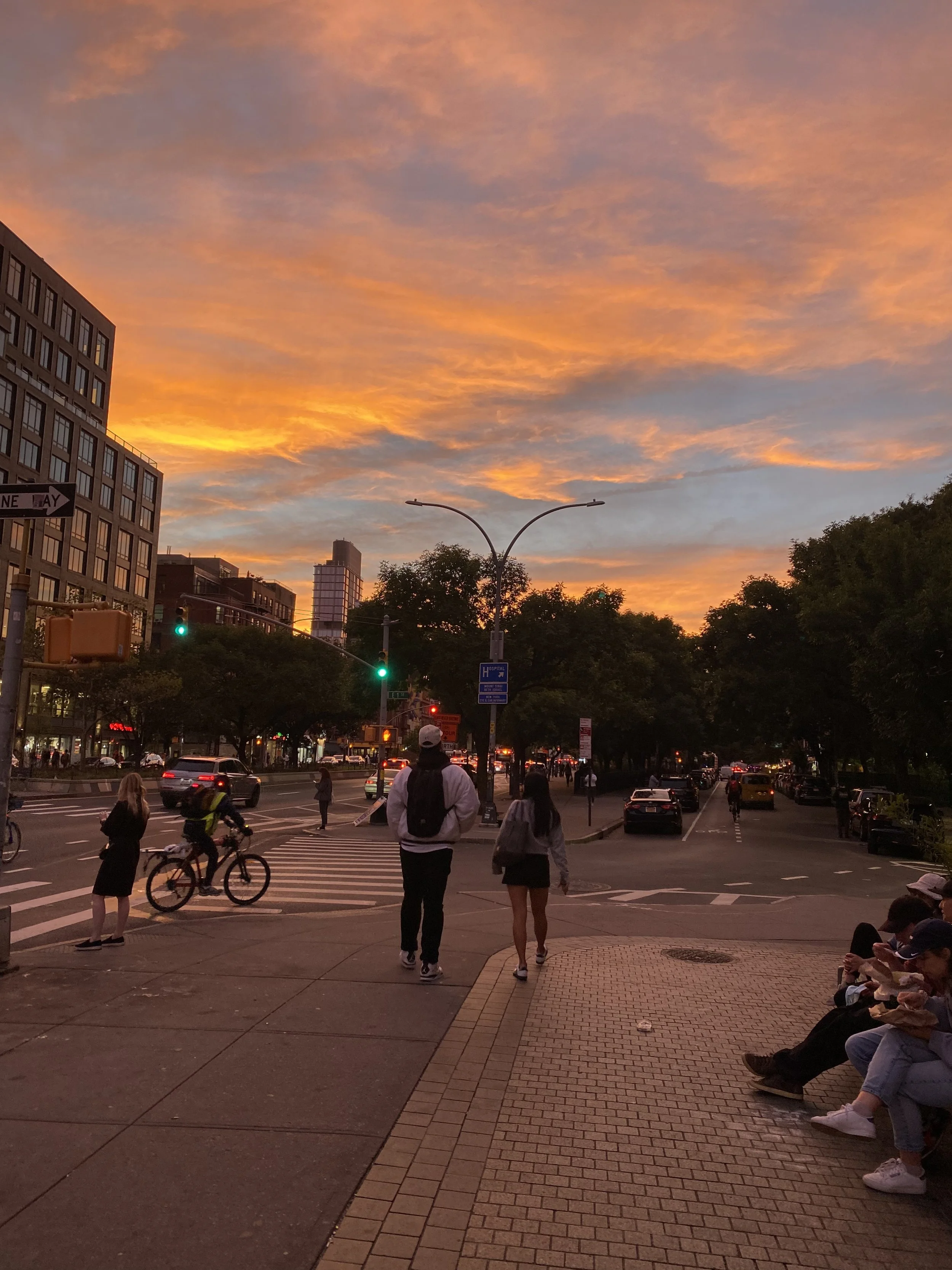 A city street at sunset with a colorful sky, people walking, and a cyclist on the crosswalk.