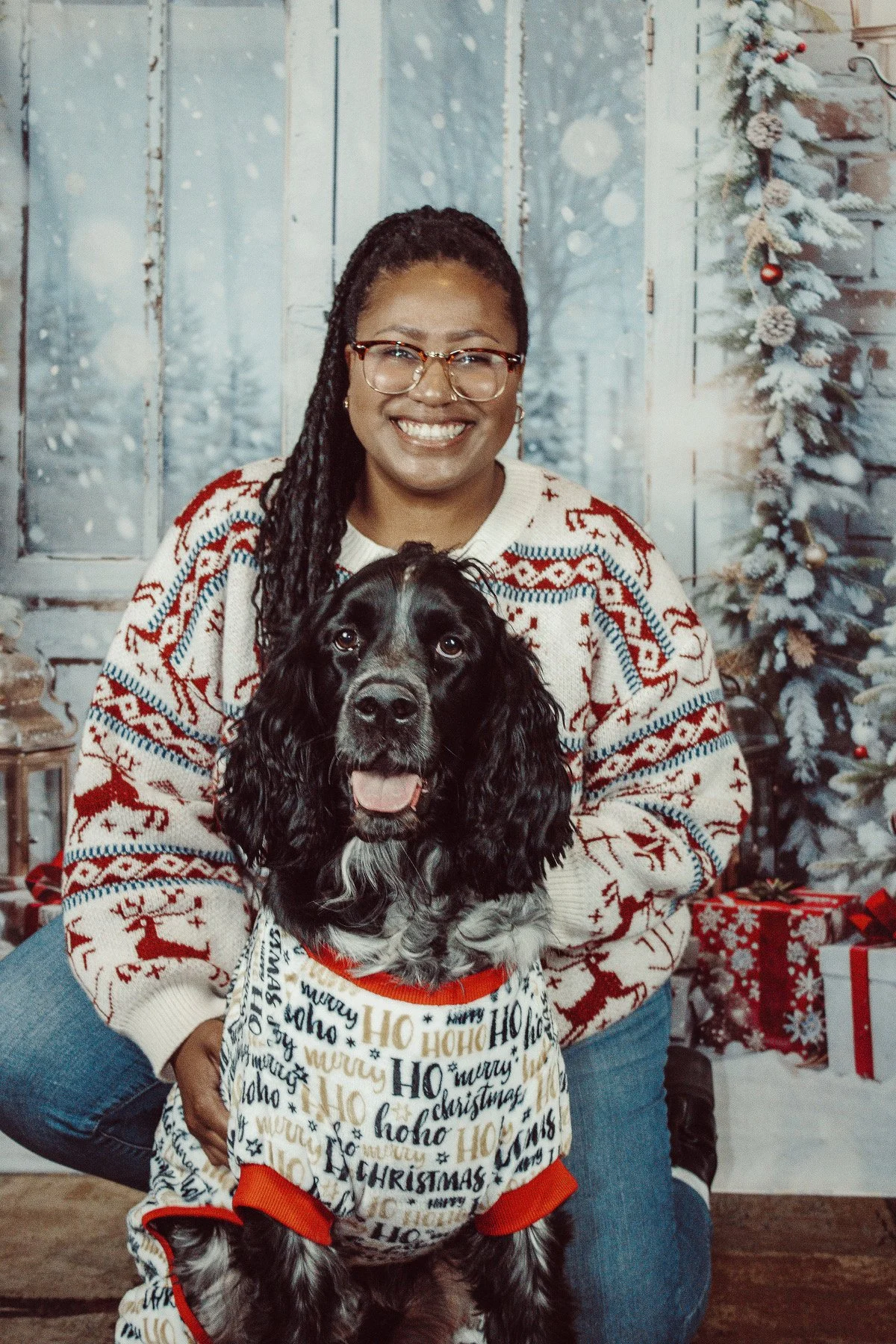 A woman with glasses and braided hair smiling while holding a black and white dog wearing a Christmas-themed sweater. They are in a festive setting with a decorated Christmas tree, snowflakes on the window, and wrapped gifts in the background.